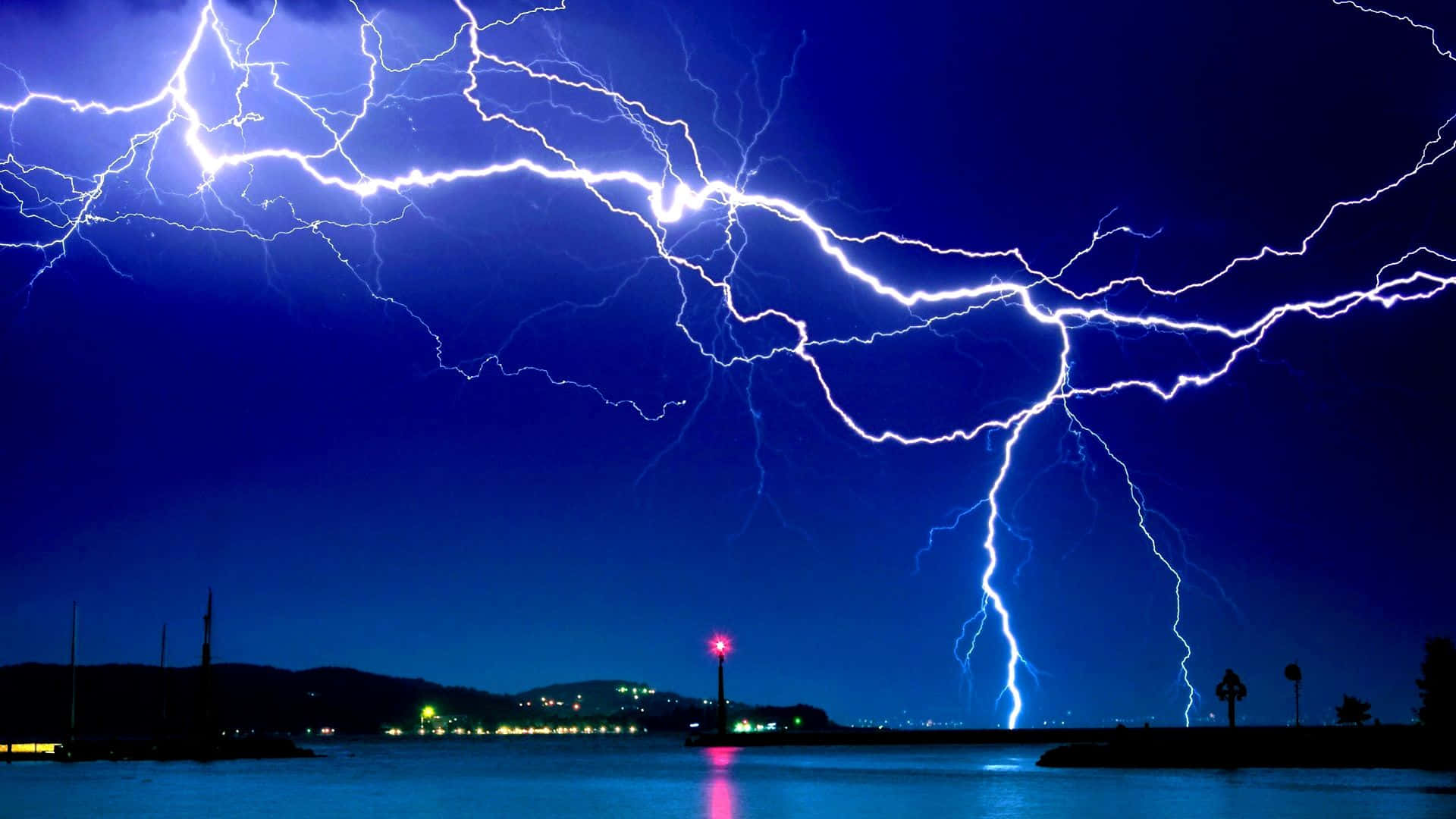 美麗的閃電劃破清澈夜空 壁紙 — 戲劇性的夜空被鮮豔的藍色閃電劈開，平靜的水面上有遠處的城市燈光和發光的燈塔。