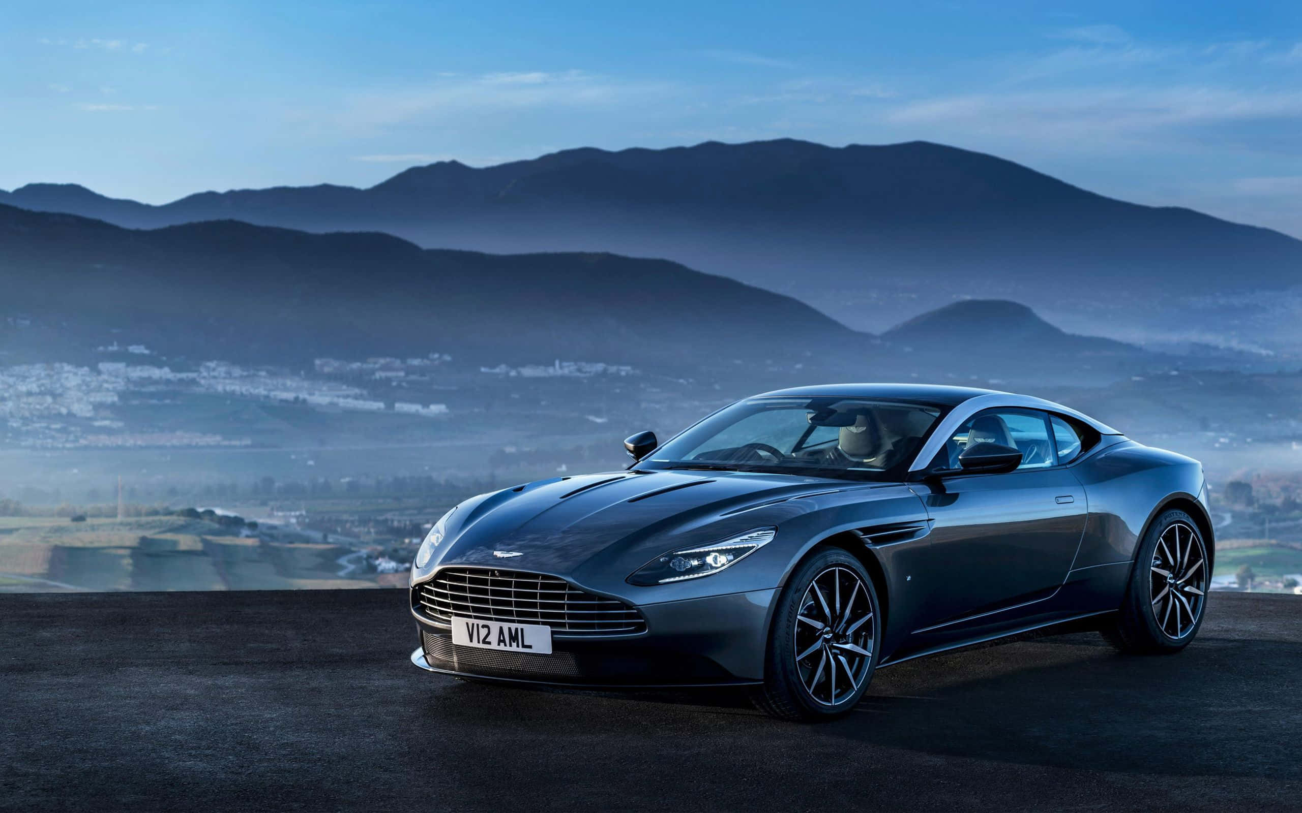 「美麗的阿斯頓·馬丁 Superleggera」 壁紙 — 一架時尚的深灰色Aston Martin跑車停在懸崖上，俯瞰著黎明時分霧氣瀰漫的山脈和山谷小鎮。
