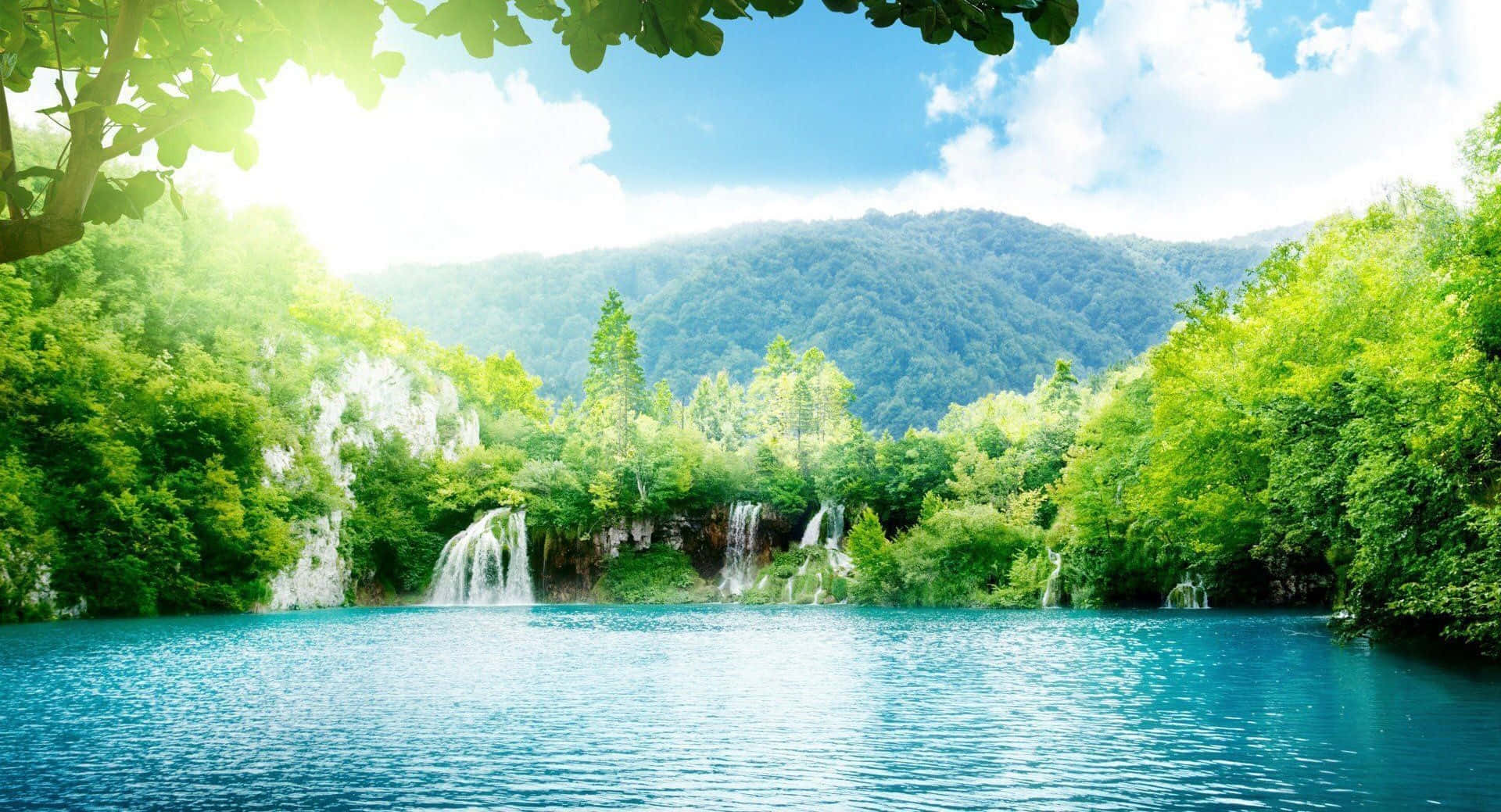 美麗的陽光湖與瀑布 壁紙 — 陽光穿過茂密的綠樹，框住一個寧靜的碧綠湖泊，背景有層疊的瀑布和森林覆蓋的山脈。