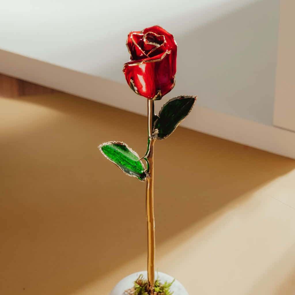 美麗紅色人造玫瑰花攝影畫 壁紙 — 一朵紅玻璃玫瑰，金色莖和綠葉，優雅地立在白色花瓶裡，放在溫暖的木地板上。