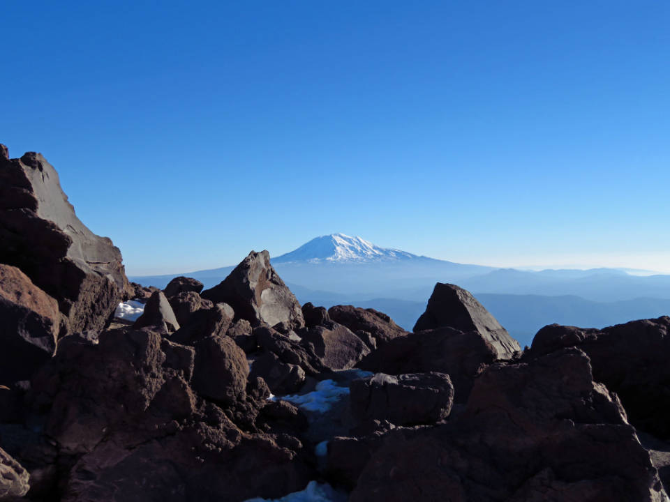 美麗藍色山峰 壁紙 — 一座白雪覆蓋的山峰在清澈的藍天下雄偉地聳立，前景是崎嶇的火山岩。