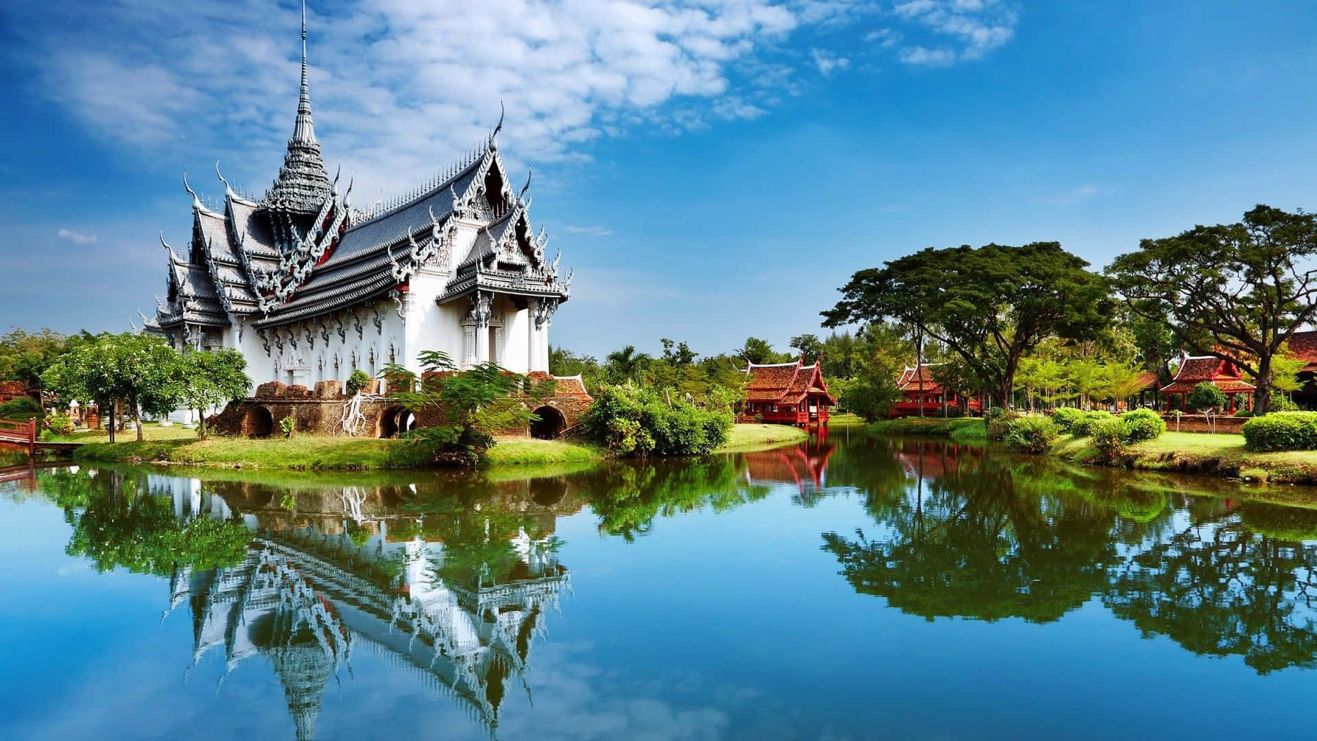 白色泰國寺廟倒映在平靜的水面上，周圍有茂密的綠樹和藍天。