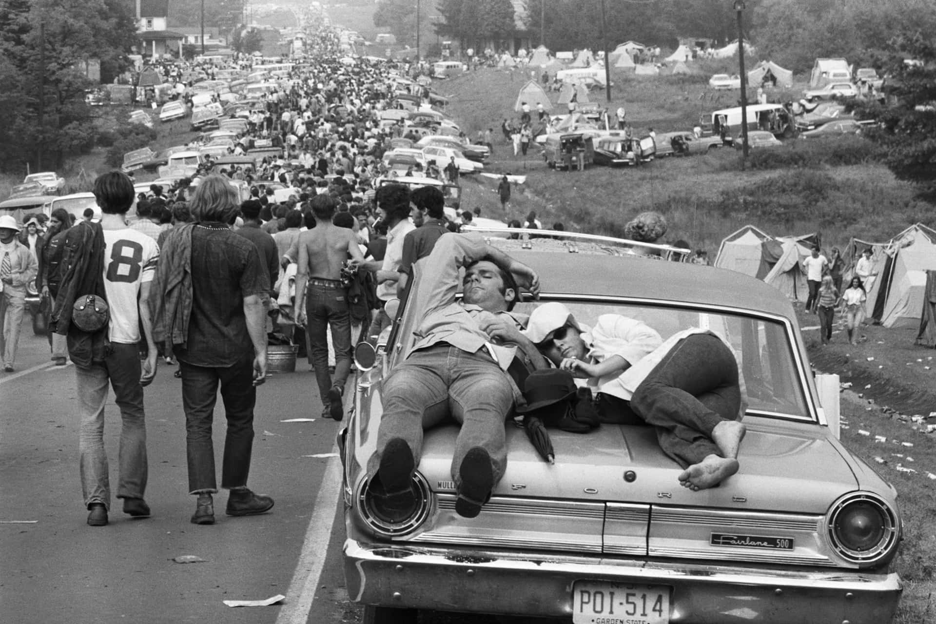 兩人在1960年代音樂節的經典汽車引擎蓋上休息，背景是人群和帳篷。