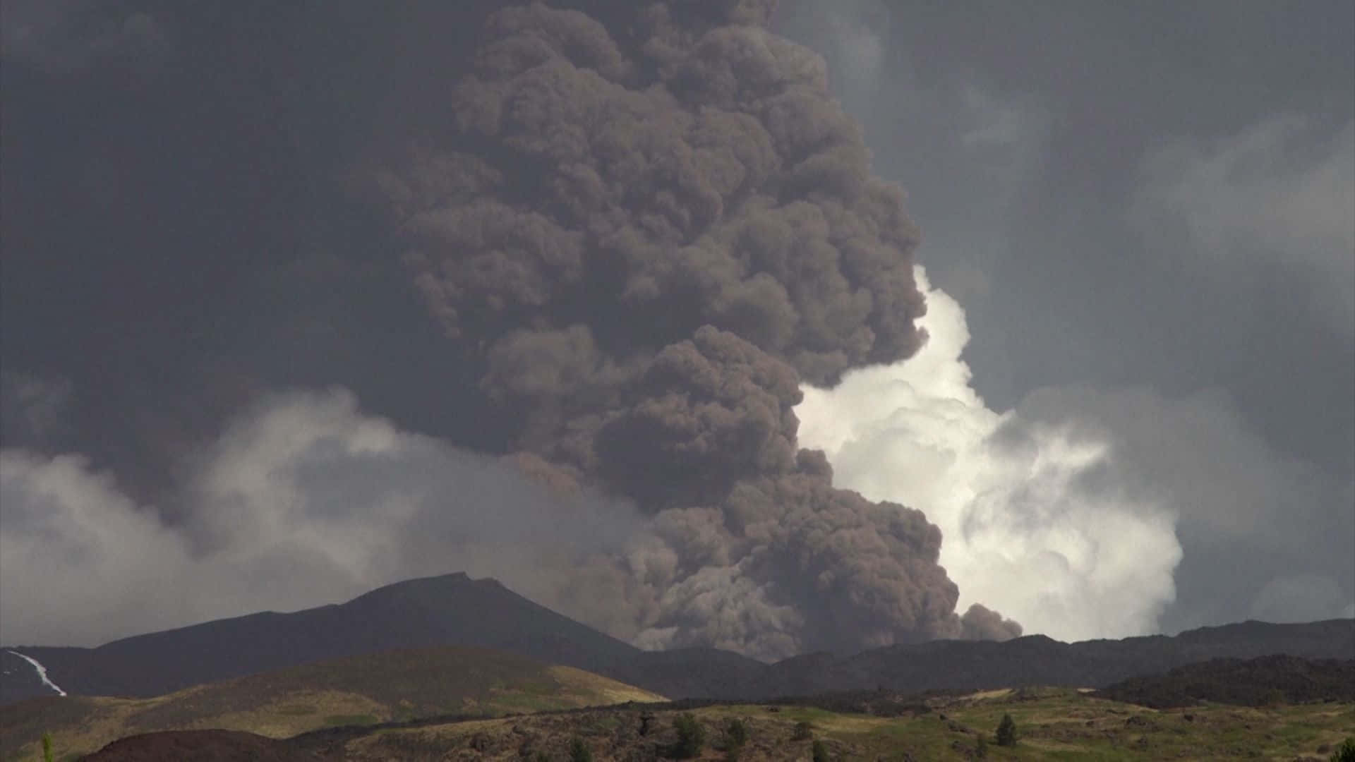 義大利埃特納火山濃黑火山灰 壁紙 — 巨大的深色火山灰柱從火山噴發而出，背景是風暴天空和翻滾的雲層。