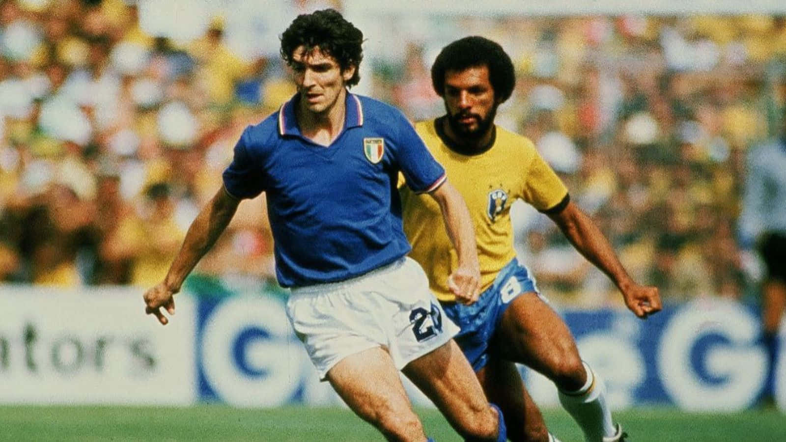 義大利足球傳奇Paolo Rossi 壁紙 — 兩名足球運動員在陽光明媚的球場上激烈競爭，背景是模糊的人群。