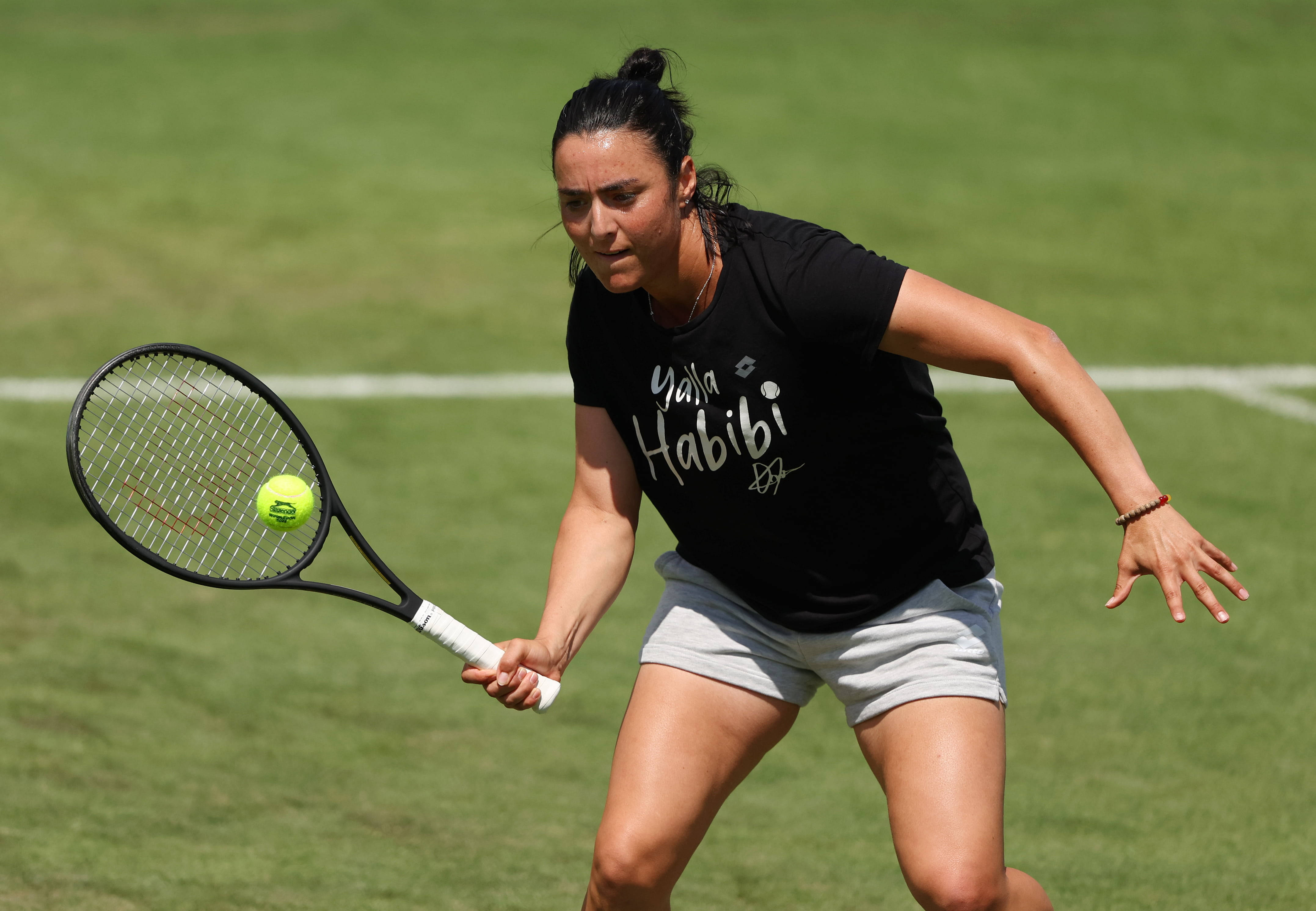 翁斯·贾贝爾展示「Yalla Habibi」運動衫 壁紙 — 一位專注的女網球手在草地場上向前撲出，用球拍擊打一個亮黃色的球。