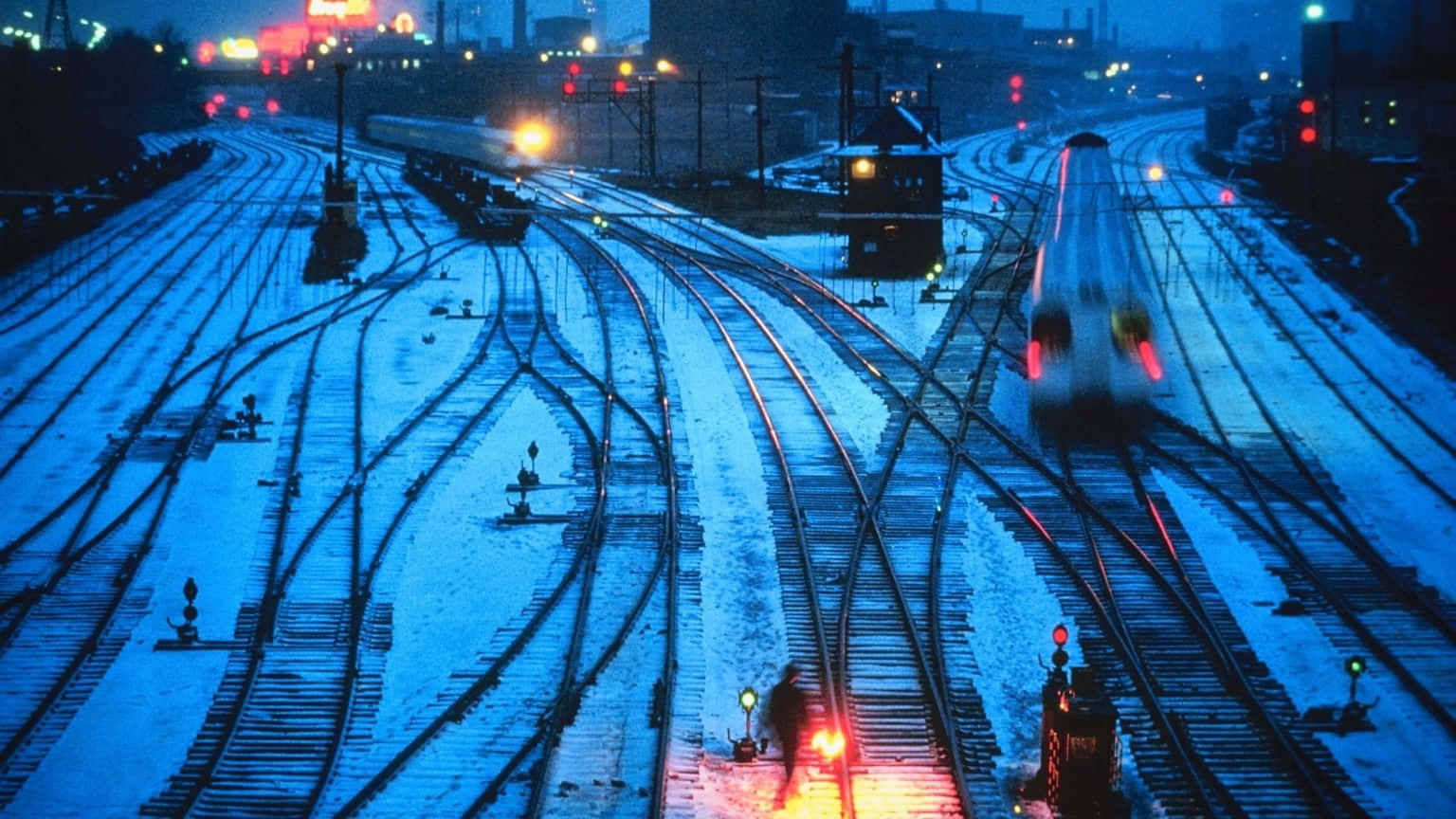 夜晚被雪覆蓋的火車軌道，有移動的火車和發光的信號燈。