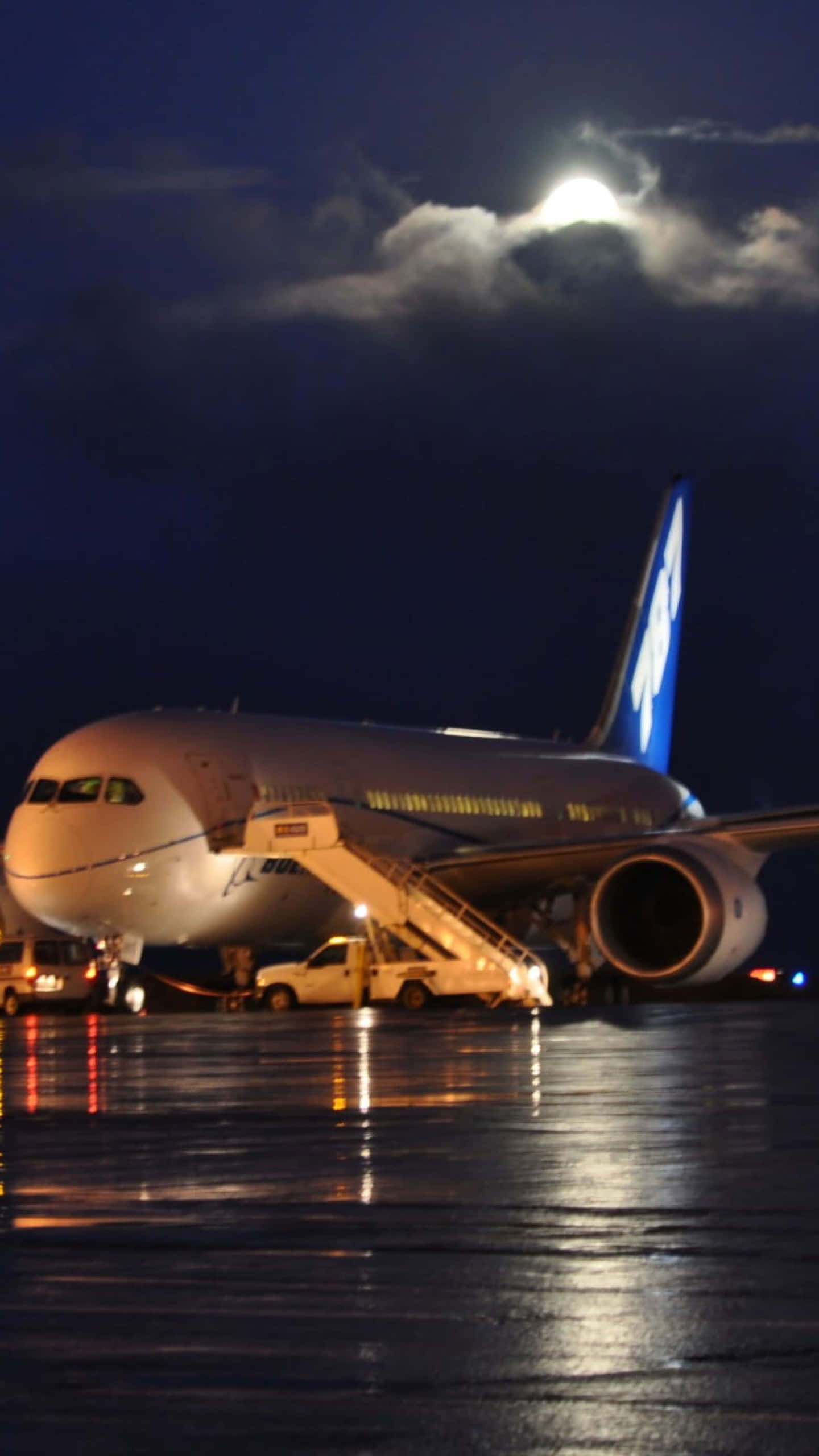 翱翔天際 壁紙 — 一架大型飛機在夜晚停放在濕滑的停機坪上，明亮的月亮和雲朵下，地面燈光反射在路面上。