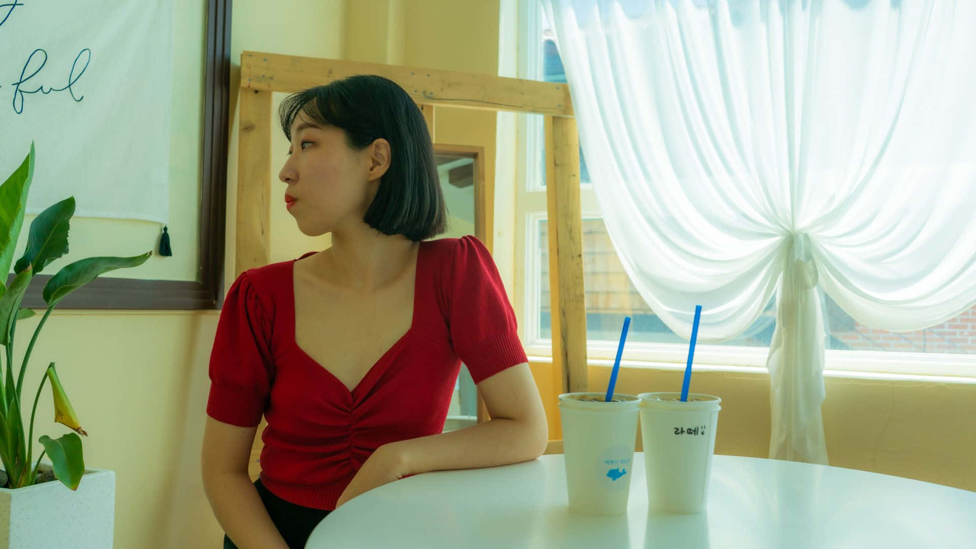 耀眼K-pop偶像登台演出 壁紙 — 一位身穿紅色上衣的女性坐在窗邊的白色桌子旁，窗簾是透明的，旁邊有兩杯飲料。