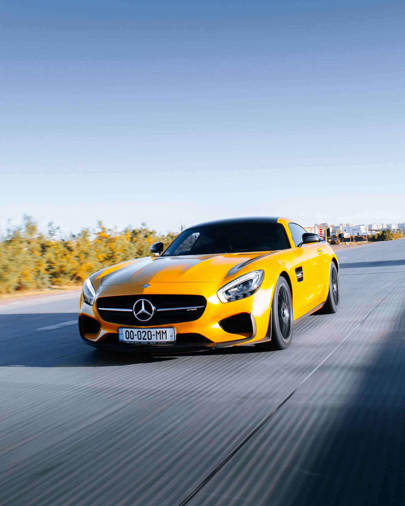 黃色Mercedes-AMG GT S在道路上疾馳，背景有秋天的樹木和城市天際線。