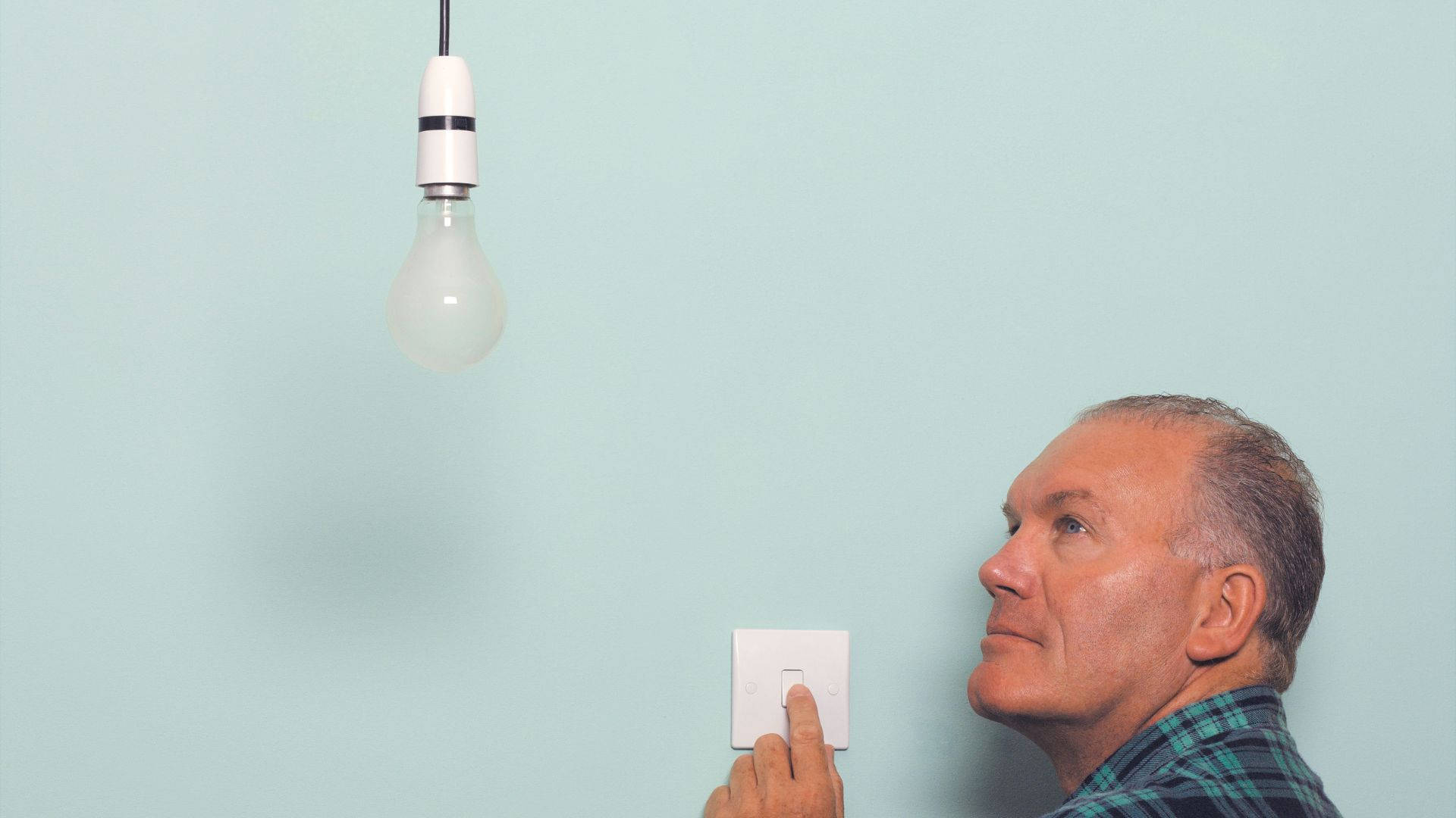 老先生檢查燈泡開關 壁紙 — 一個穿著格仔恤衫的男人按下電燈開關，同時抬頭望向掛在淺藍色牆上的一個未亮起的燈泡。
