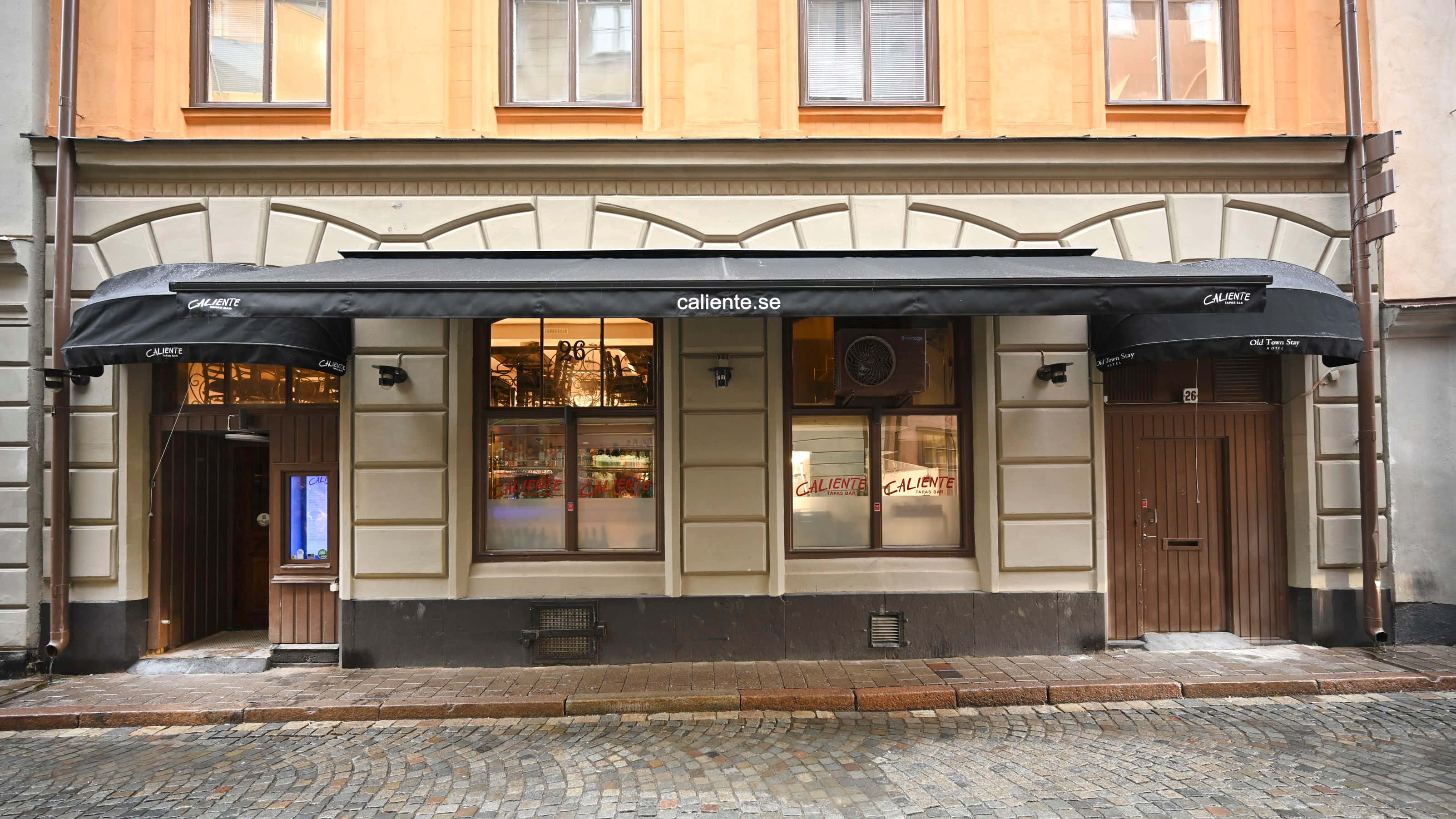 老城Callente便利店 壁紙 — 一間迷人的歐洲咖啡館，帶有黑色遮陽篷和溫暖的室內燈光，坐落於鵝卵石街道上。