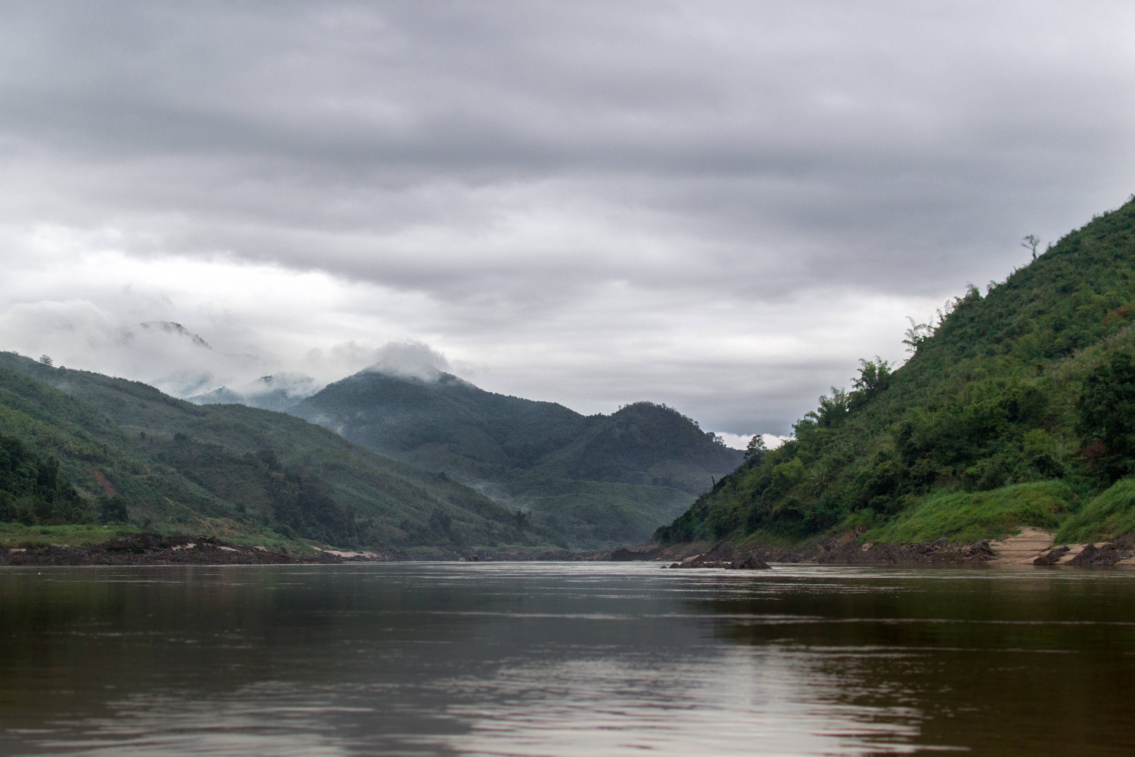 老撾湄公河浩瀚河景 壁紙 — 一條平靜的河流蜿蜒穿過鬱鬱蔥蔥的青山，天空陰沉，薄霧籠罩著遠峰。