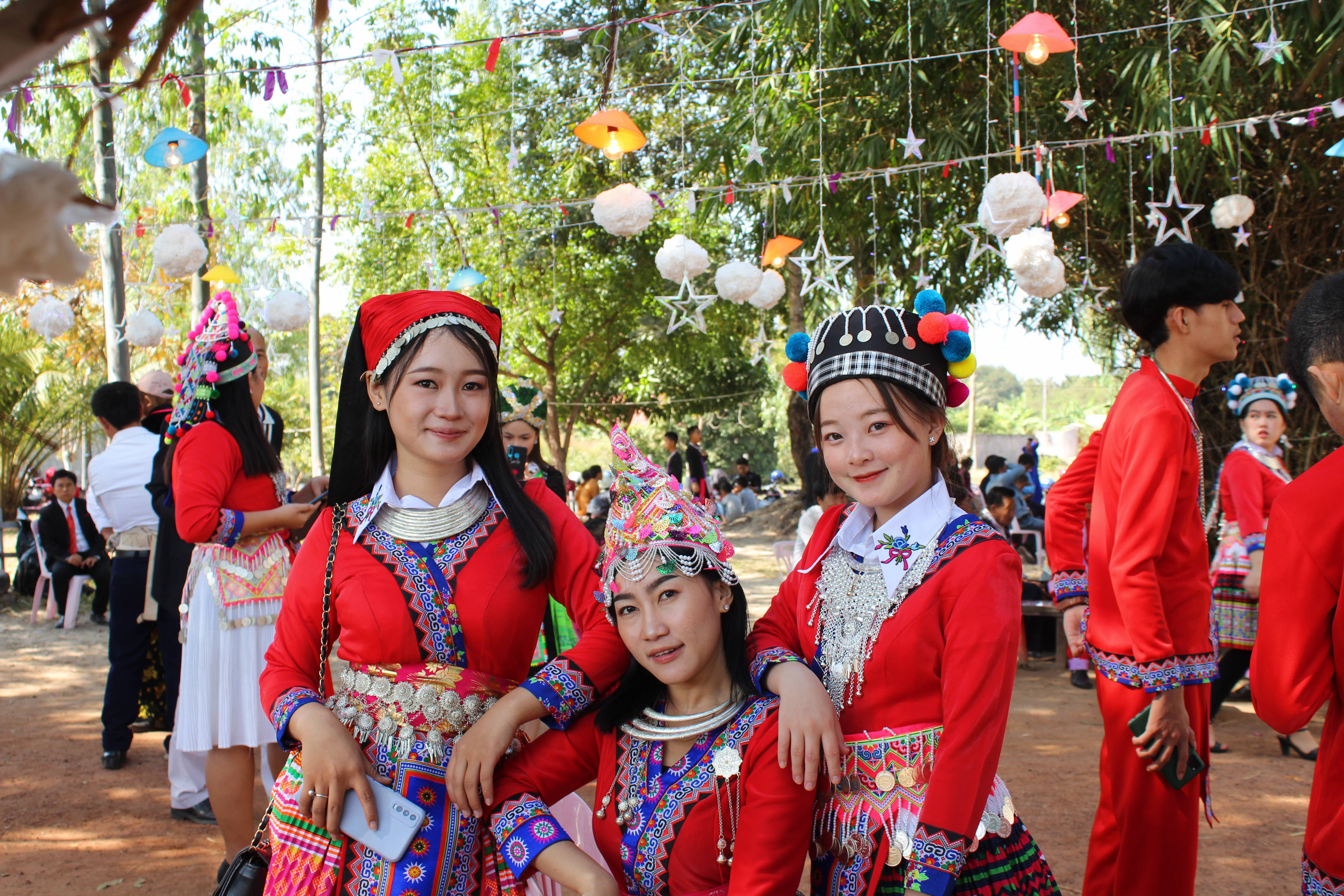 老撾穿傳統服飾少女 壁紙 — 三位年輕女性身穿鮮豔的傳統紅色服飾，佩戴精緻的銀飾和色彩繽紛的頭飾，在陽光明媚的戶外聚會中，於節日燈光下溫暖地微笑著看向鏡頭。