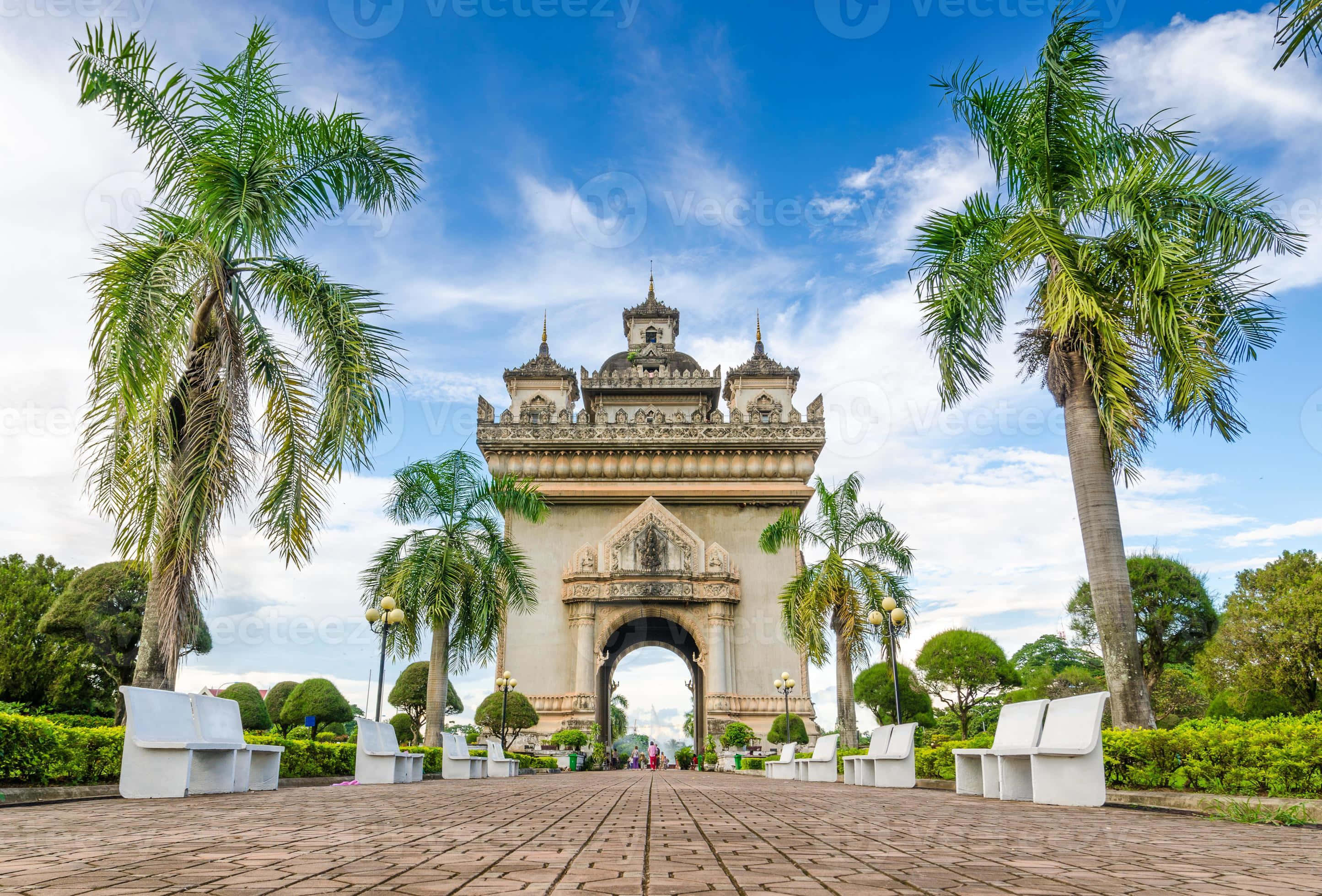 老撾萬象巴特寮紀念碑的壯麗全景 壁紙 — 一個宏偉華麗的石拱門矗立在明亮的藍天下，兩旁是高聳的棕櫚樹和白色長椅，位於鋪砌的人行道上。