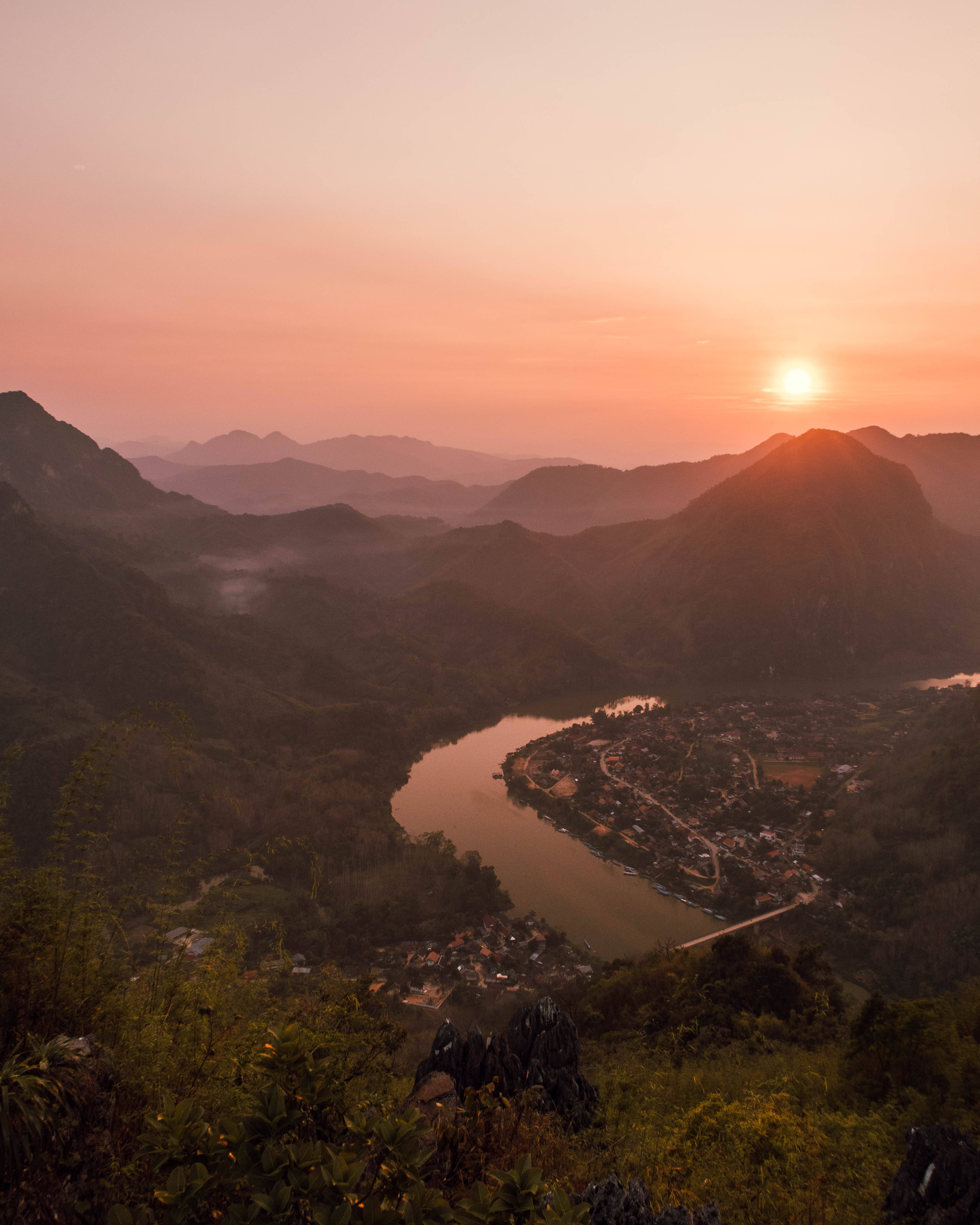 老撾農基亞河 壁紙 — 寧靜的夕陽將蜿蜒的河流和依偎的村莊沐浴在溫暖的橙色和粉紅色調中，周圍是霧氣繚繞的山巒。