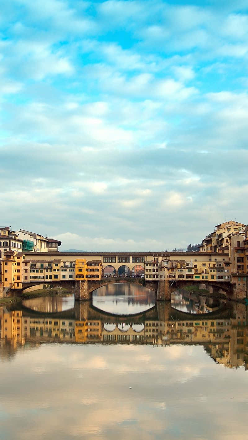 老橋獨特景觀：秋日河面倒影 壁紙 — 佛羅倫斯歷史悠久的舊橋（Ponte Vecchio）在平靜的阿諾河上美麗地倒映，柔和的藍天中散佈著雲朵。