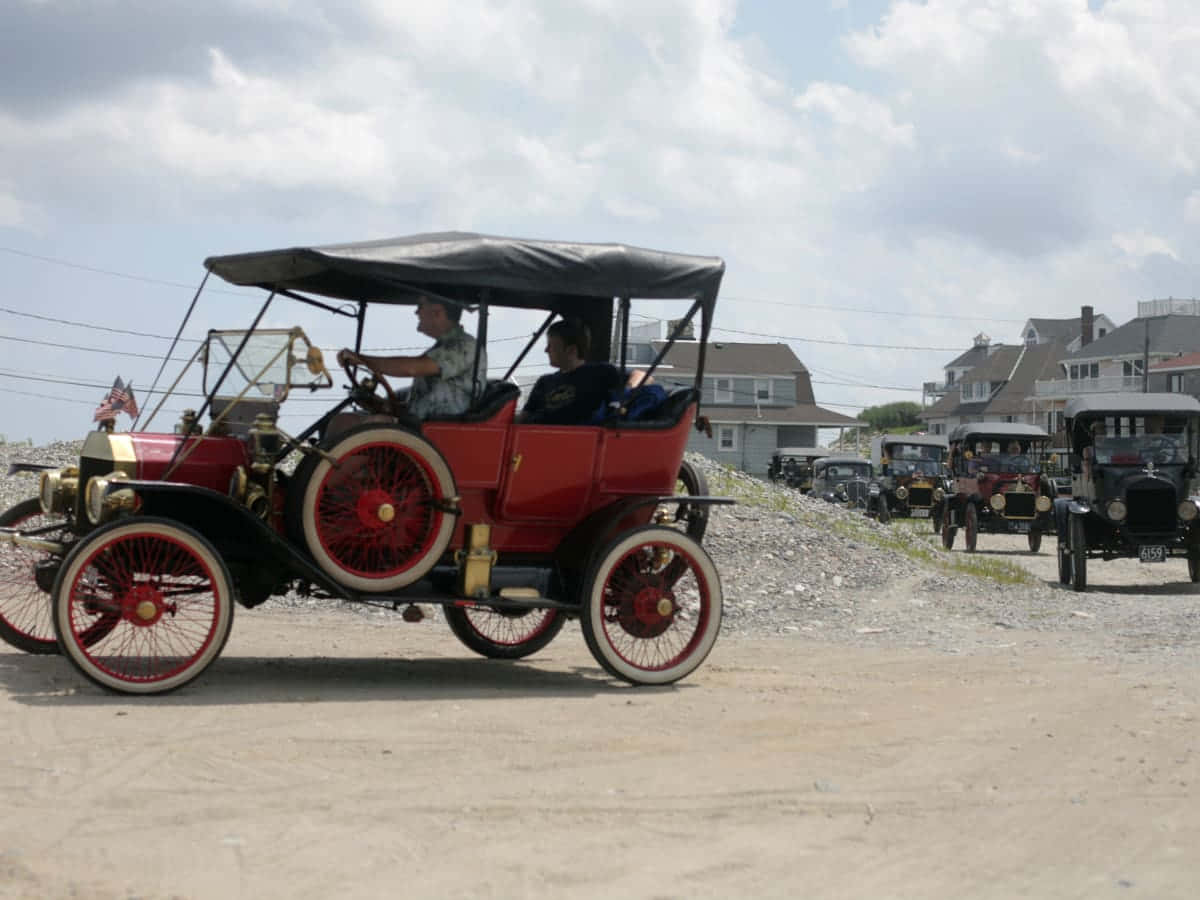 一輛復古紅色福特Model T，有兩個人駕駛在碎石路上，背景有其他經典汽車。