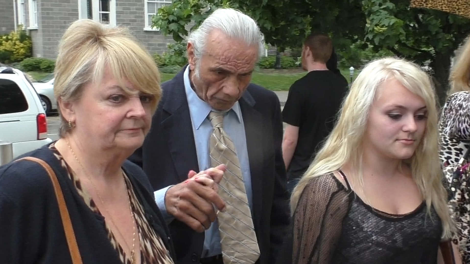一位長者男士在戶外與兩位女士手牽手，旁邊有一間房屋和一輛停泊的汽車。