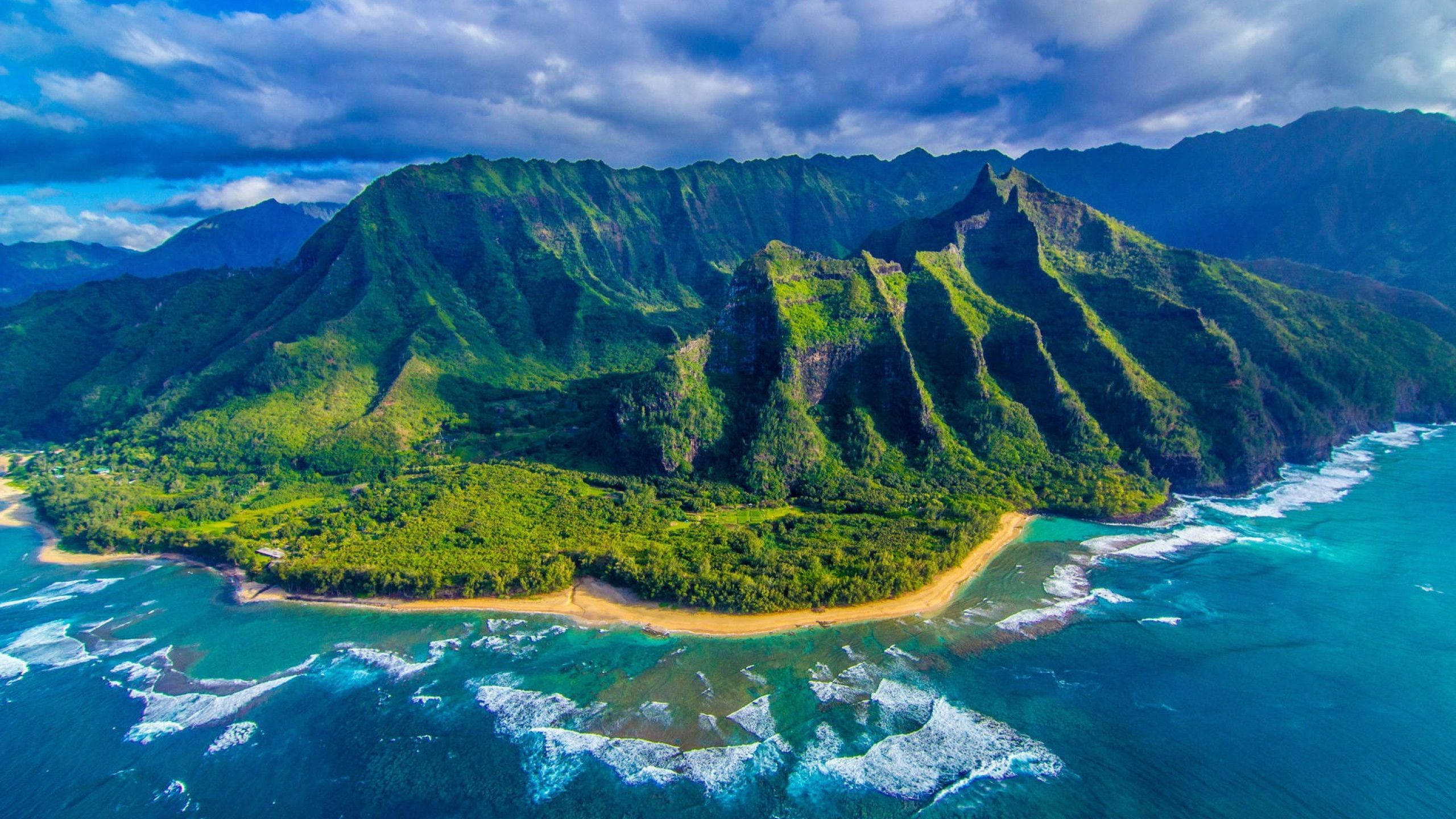 考艾島 壁紙 — 翠綠山巒與碧綠海洋交匯的鳥瞰圖，沙灘和拍岸浪花在戲劇性的雲層下。