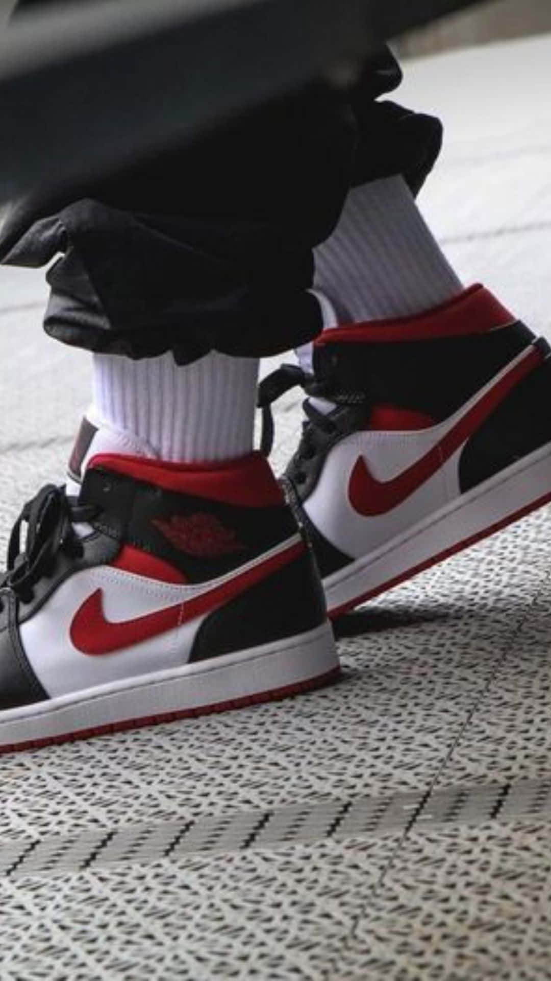 耐克 Jordan Air 1 男鞋 壁紙 — 黑色、紅色和白色Jordan 1波鞋嘅特寫，配白色襪同黑色褲，企喺有圖案嘅行人路上。