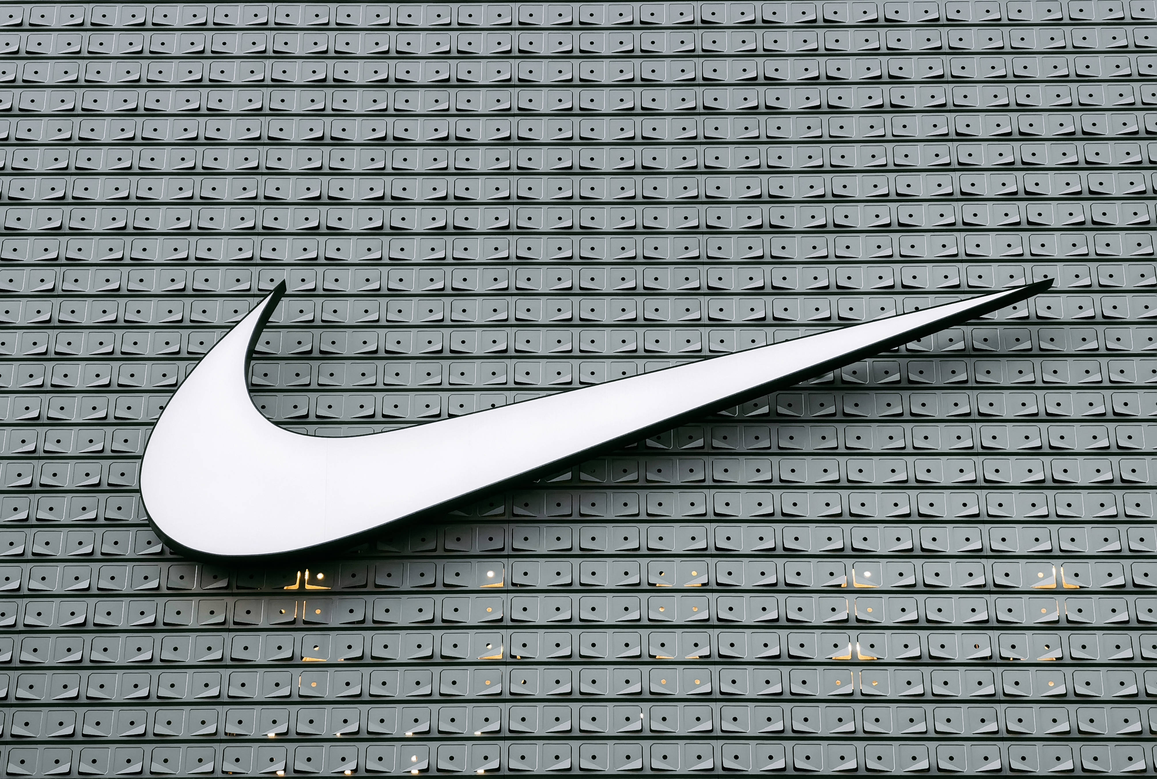 耐克勾形標誌廣告牌 壁紙 — 一個大型白色Nike swoosh標誌安裝在帶有重複方形圖案的灰色紋理牆上。
