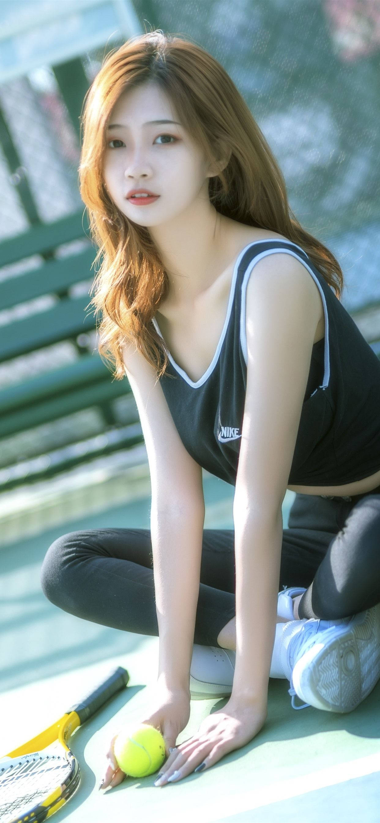 耐克女孩模特持網球拍 壁紙 — 一位身穿黑色背心的年輕女子跪在網球場上，手持黃色球，旁邊放著球拍。