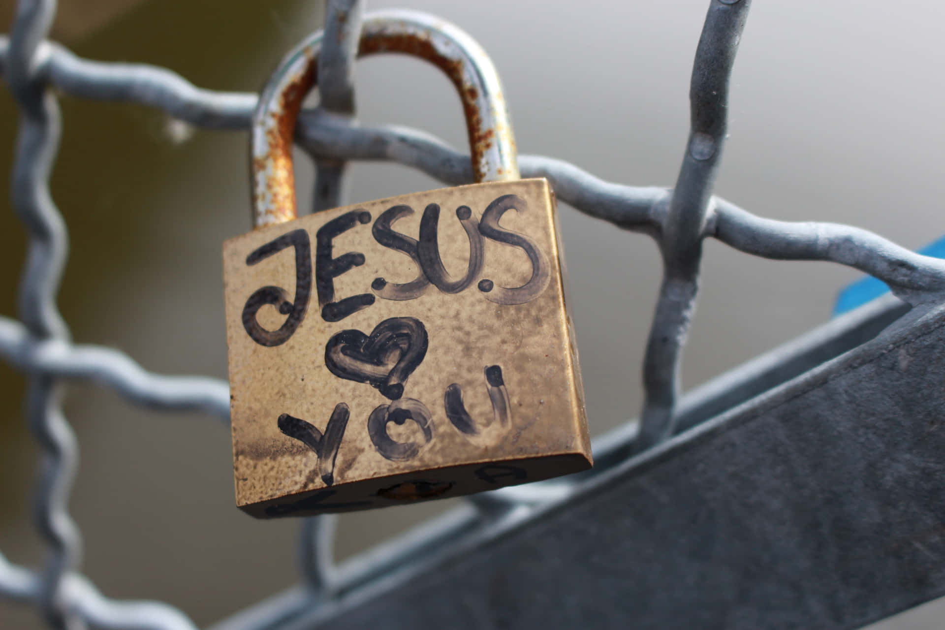 一個青銅掛鎖，上面寫著「JESUS ❤️ YOU」，掛在鐵絲網圍欄上。