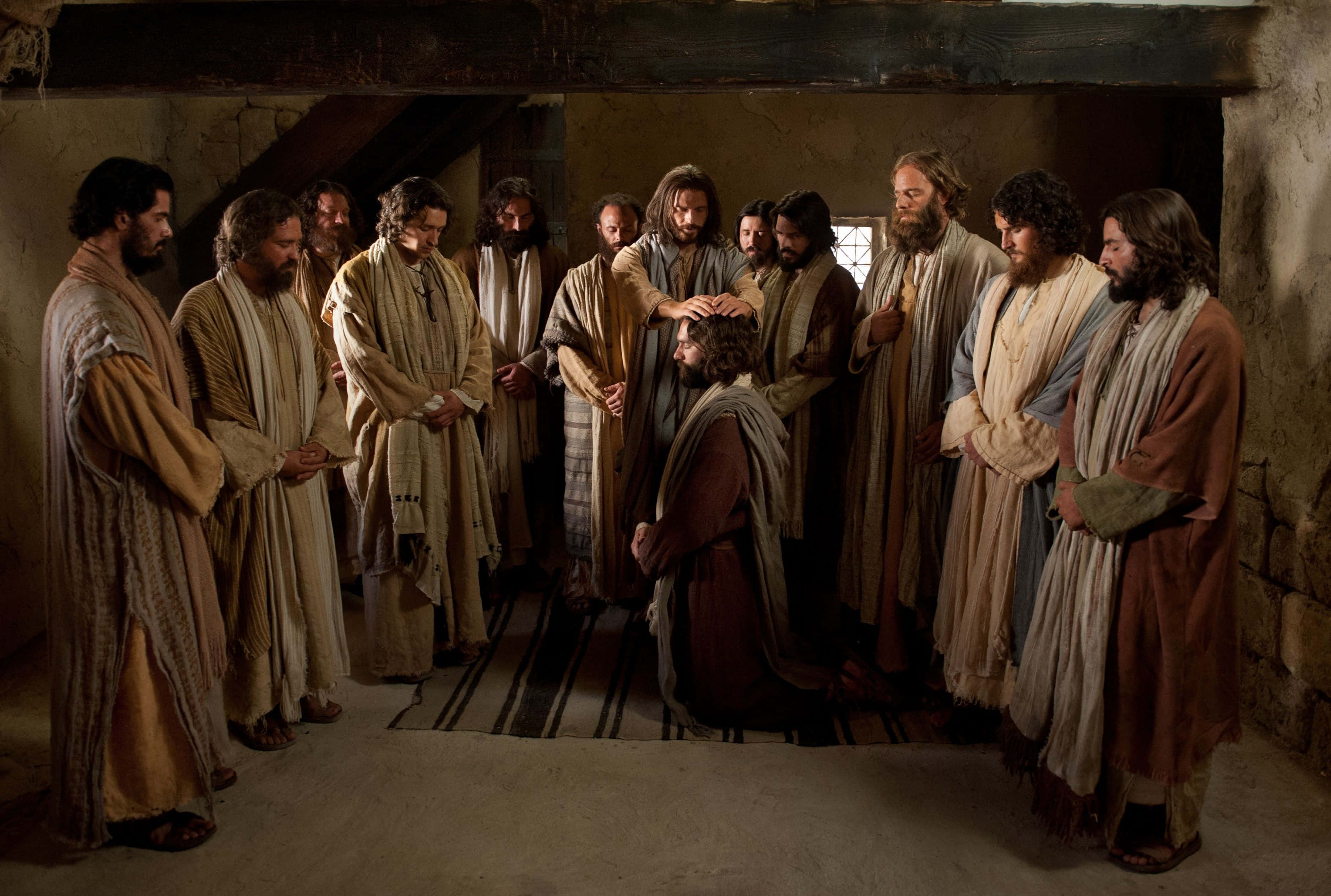 Jesus Disciples 桌布 壁紙 — 耶穌祝福一位跪下的門徒，祂的門徒聚集在一個質樸的石室中，有木樑和柔和自然光。