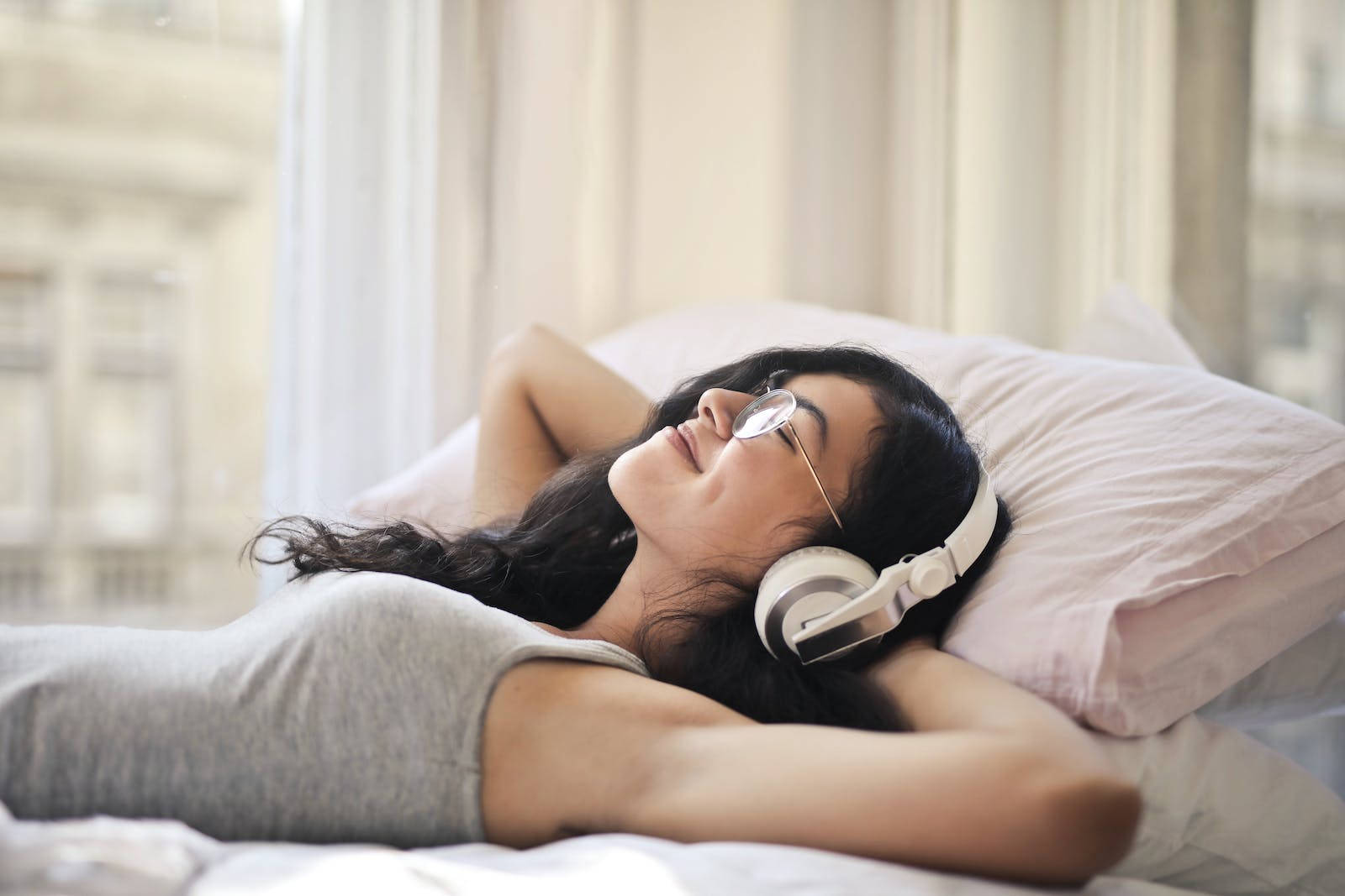 聆聽放鬆音樂 壁紙 — 一位戴著眼鏡和耳機的女性在明亮的窗邊，在柔軟枕頭的床上平靜地休息。