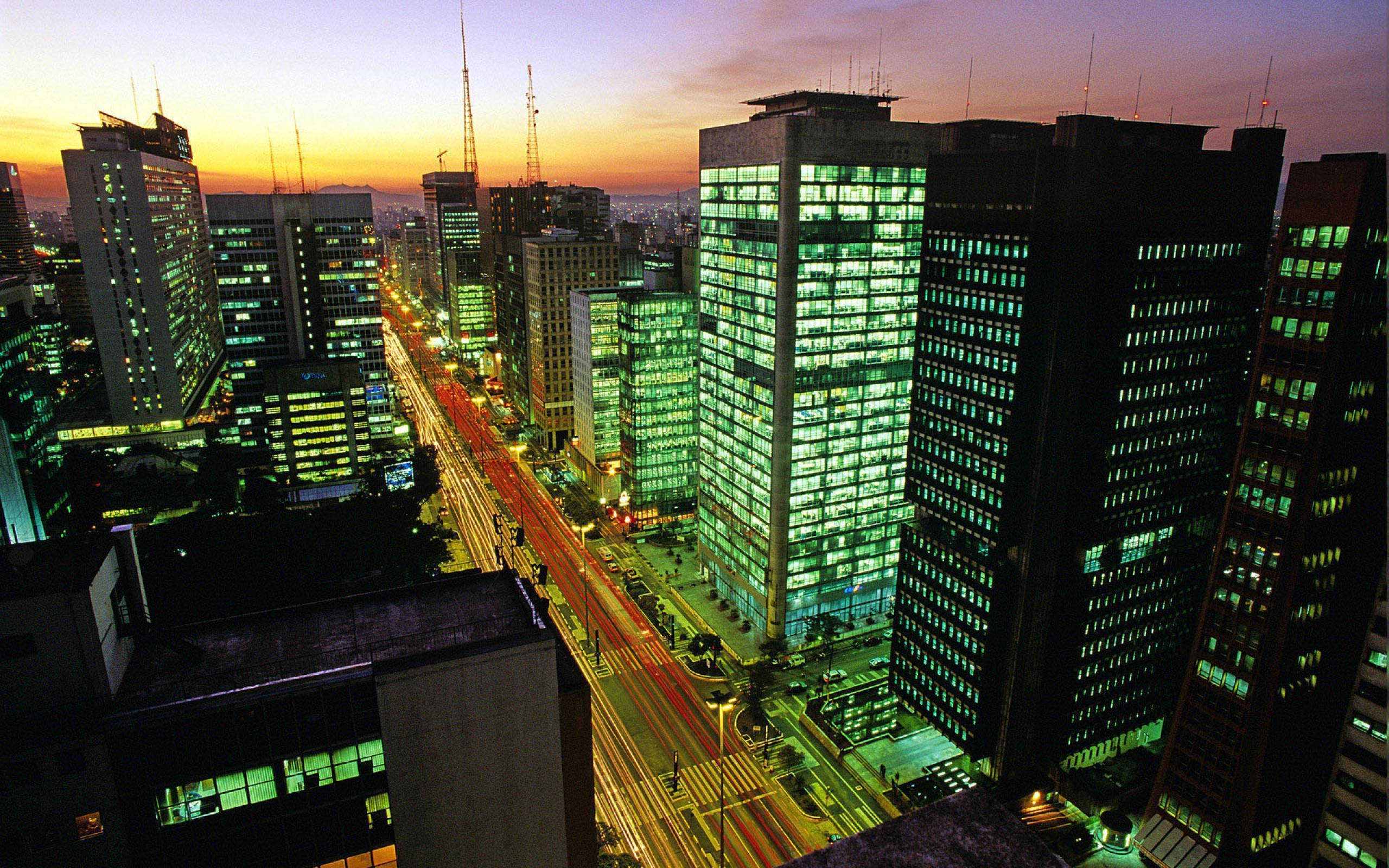 聖保羅巴西綠色城市燈光 壁紙 — 黃昏時分充滿活力的城市景觀，高聳的摩天大樓閃爍著綠光，車流的燈光劃過城市峽谷。