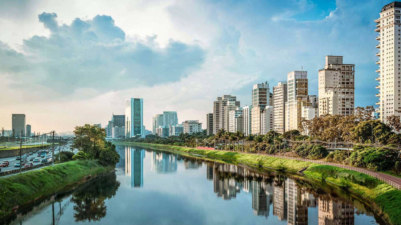 聖保羅巴西風景 壁紙 — 一條寧靜的城市河流在部分多雲的天空下，倒映著高聳的摩天大樓和鬱鬱蔥蔥的綠色河岸。