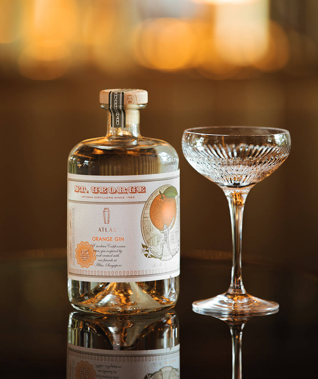 深色表面上的 St George Atlas Orange Gin 酒瓶和 coupe 杯，帶有溫暖燈光。