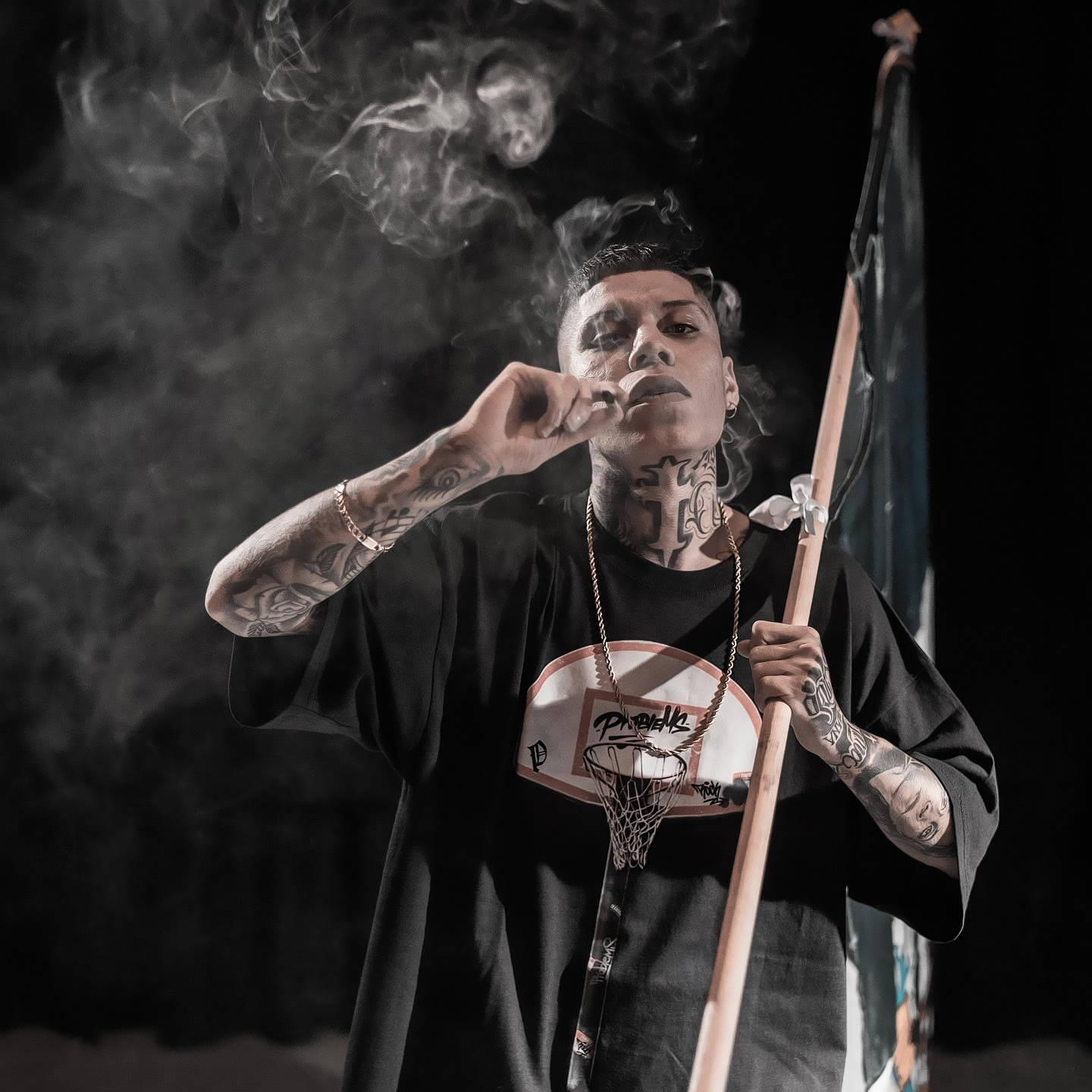 有紋身的男人抽煙，在煙霧瀰漫的黑暗環境中拿著旗幟和籃球框吊飾。