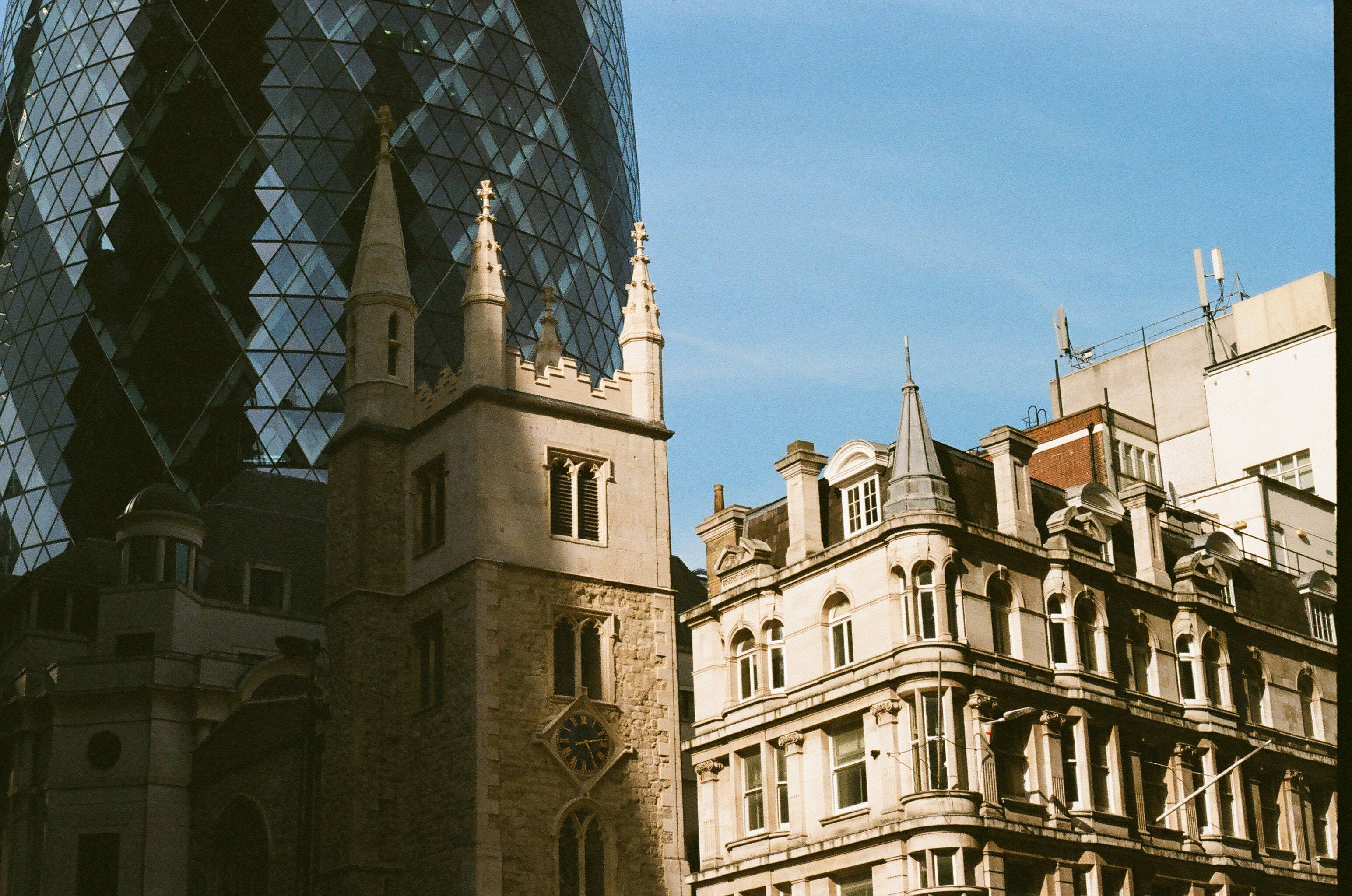 聖安德魯教堂與The Gherkin 壁紙 — 現代玻璃摩天大樓與歷史悠久的石教堂塔樓在晴朗的藍天下形成對比。