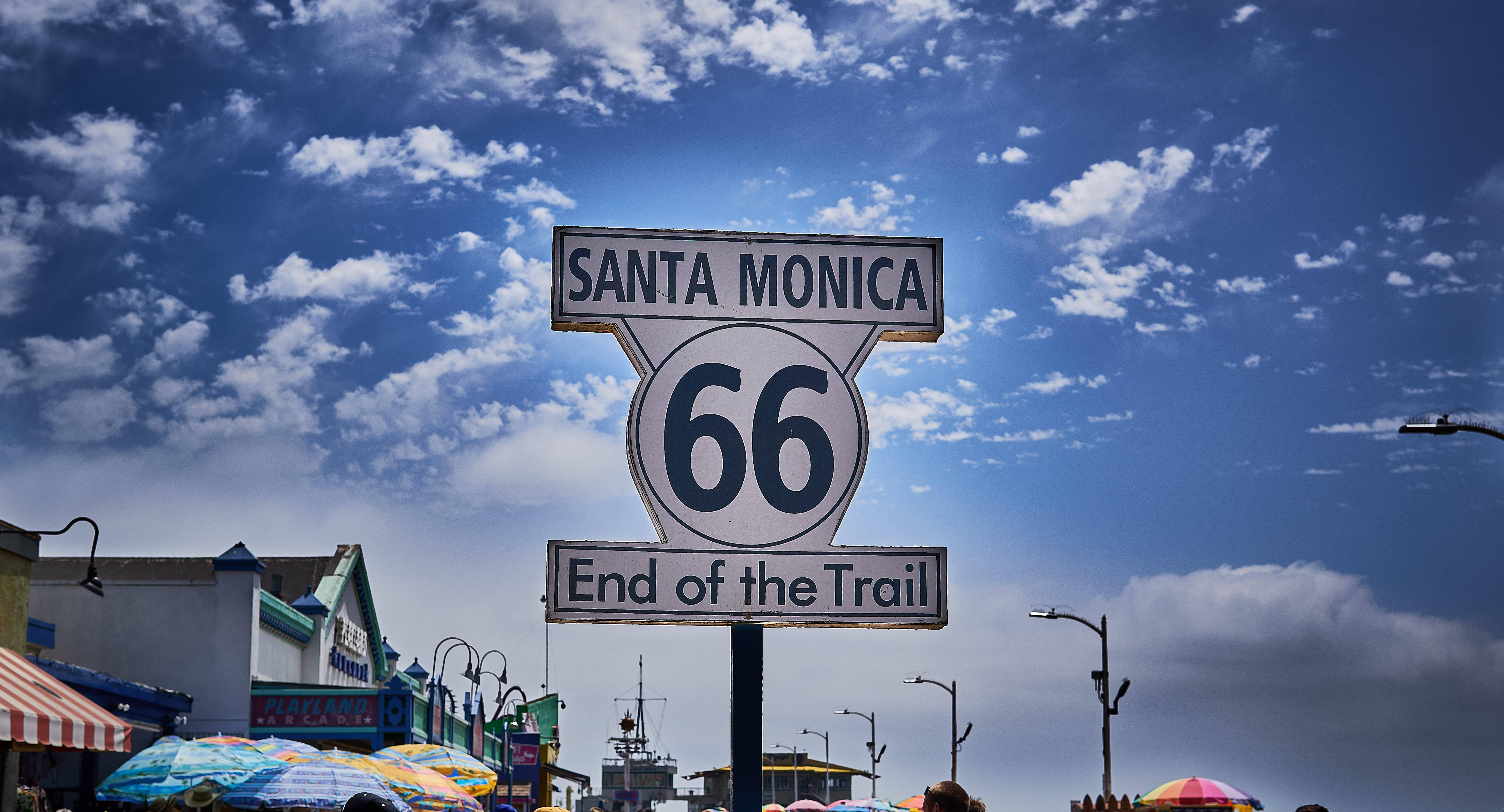 聖莫尼卡66號公路終點標誌 壁紙 — 一個寫有「Santa Monica End of the Trail」的復古66號公路標誌，矗立在藍天白雲下。