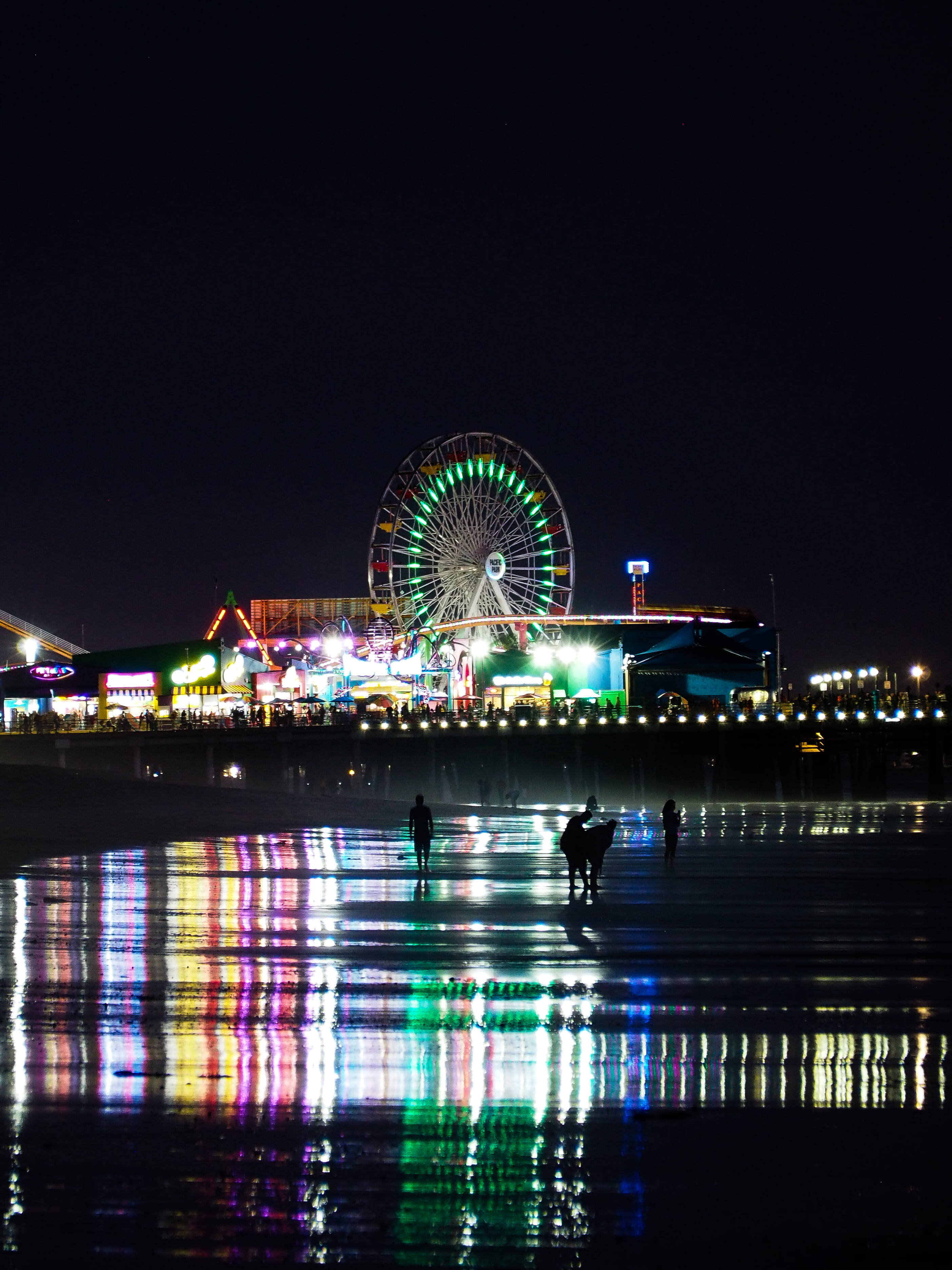 聖莫尼卡海灘彩色倒影 壁紙 — 夜晚海灘場景，充滿活力的嘉年華燈光反射在濕沙上，剪影人物在發光的摩天輪附近漫步。