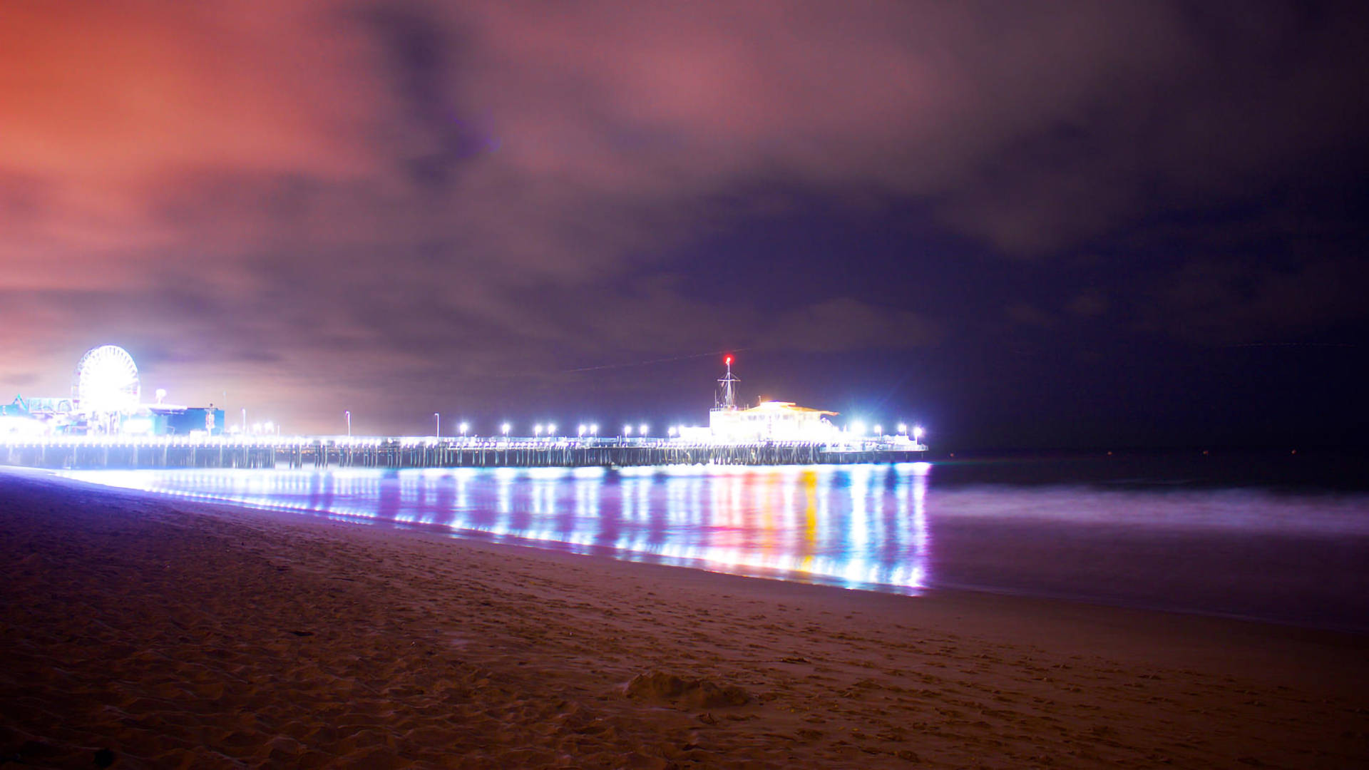聖莫尼卡璀璨燈光 壁紙 — 海灘上充滿活力的夜景，明亮的碼頭在色彩繽紛的多雲天空下延伸至海洋。