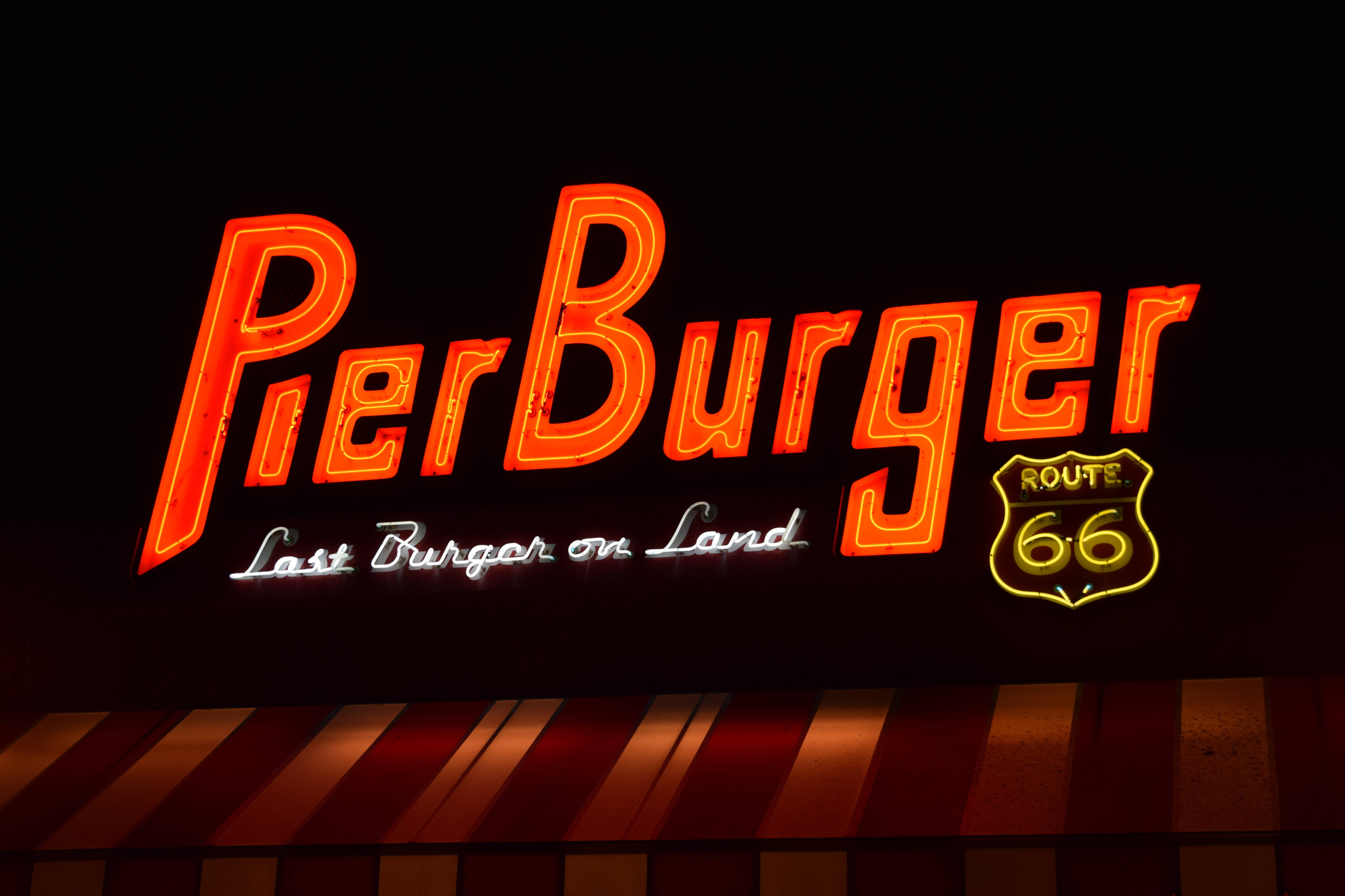 聖莫尼卡碼頭漢堡 壁紙 — 一個發光的霓虹燈招牌，上面有Pier Burger和Route 66標誌，在黑暗的夜空下，位於條紋遮陽篷上方閃耀。