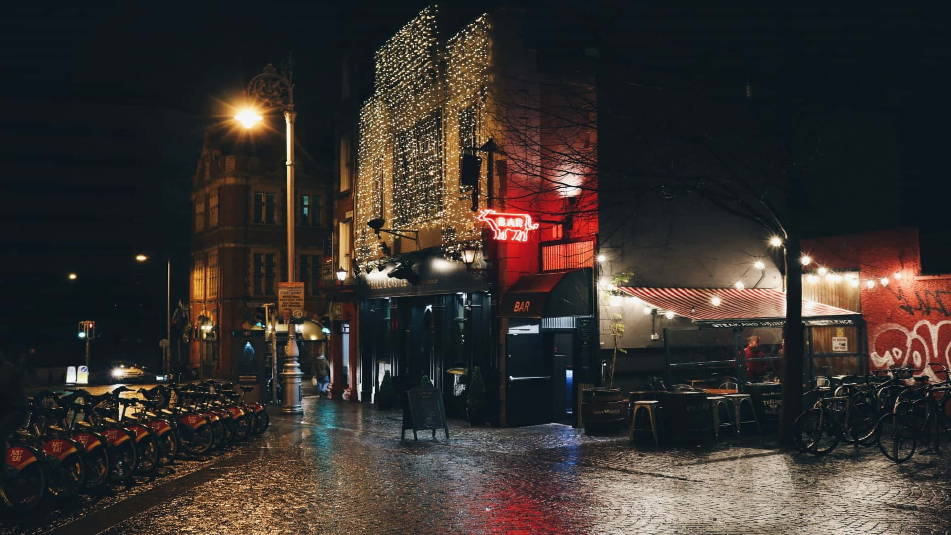 聖誕前夜雨中節日團隊背景 壁紙 — 一條濕漉漉的城市街道在夜晚被溫暖的路燈和鮮豔的霓虹燈招牌照亮，倒映在雨水浸濕的瀝青路面上。