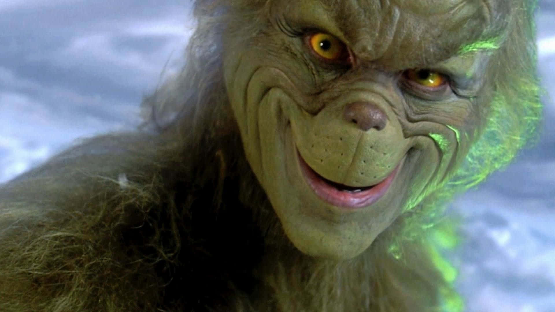 The Grinch 桌布 壁紙 — 聖誕怪傑的近距離特寫，黃色發光的眼睛和邪惡的笑容，背景是雪景。