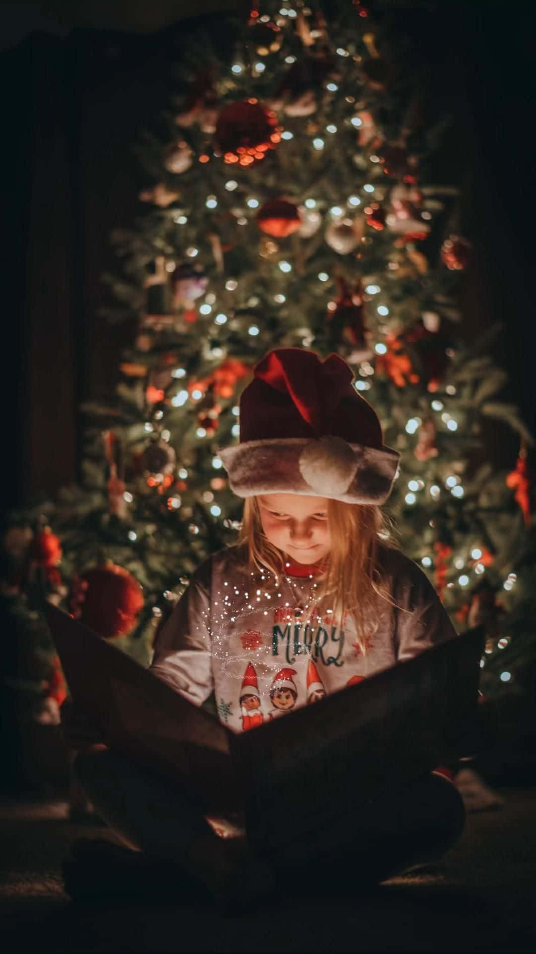 戴聖誕老人帽的小孩在亮燈的聖誕樹前讀書。