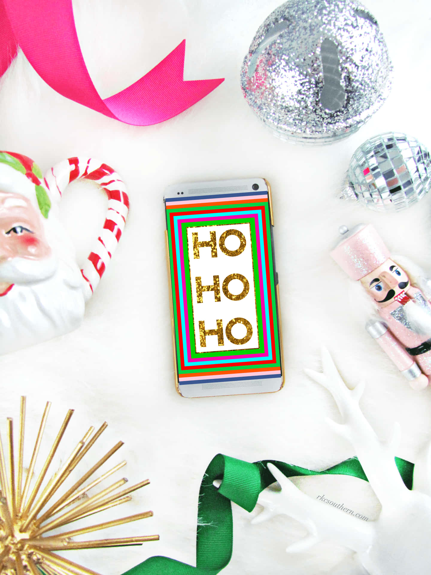 手機顯示 'HO HO HO'，被聖誕裝飾包圍，背景為白色毛絨。