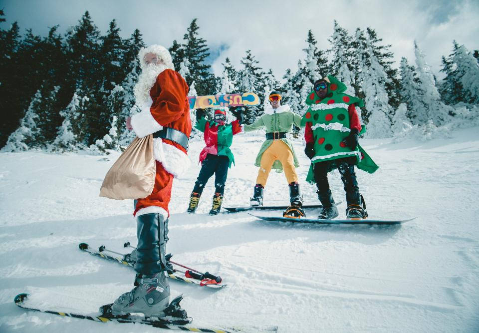 Santa Claus 桌布 壁紙 — 聖誕老人和三位穿著節日服裝的滑雪板愛好者在被常青樹環繞的雪山斜坡上擺姿勢。