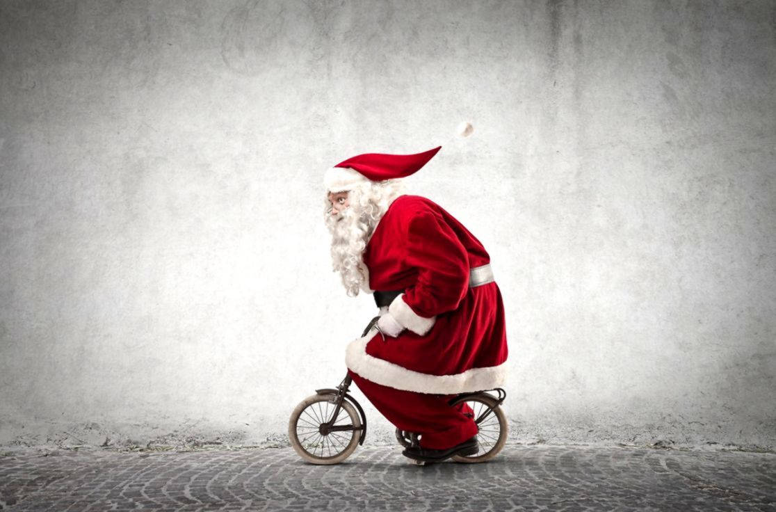 Santa Claus 桌布 壁紙 — 聖誕老人喺鵝卵石街道上，喺有紋理嘅灰色牆前面踩住一架細單車。