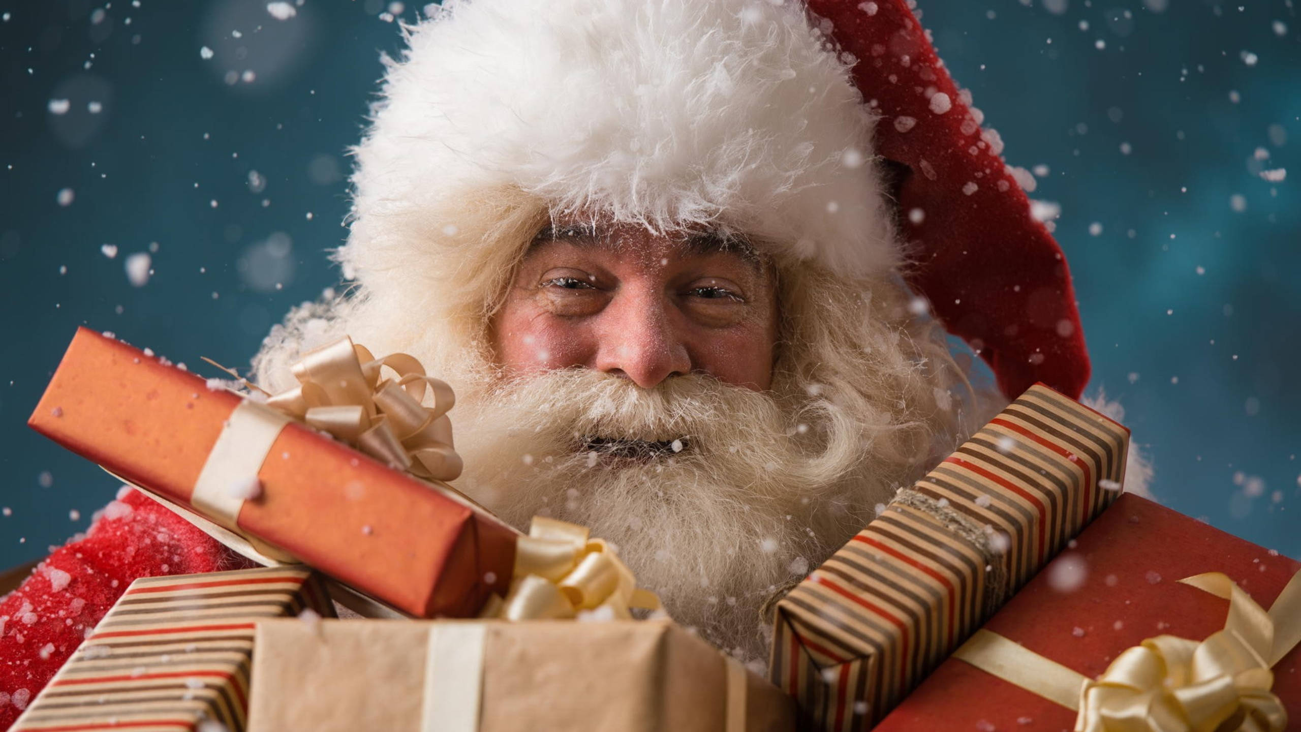 Santa Claus With Presents 壁紙 — 聖誕老人帶著溫暖的笑容從一堆包裝好的禮物後面探出頭來，雪花在深藍色背景上飄落。
