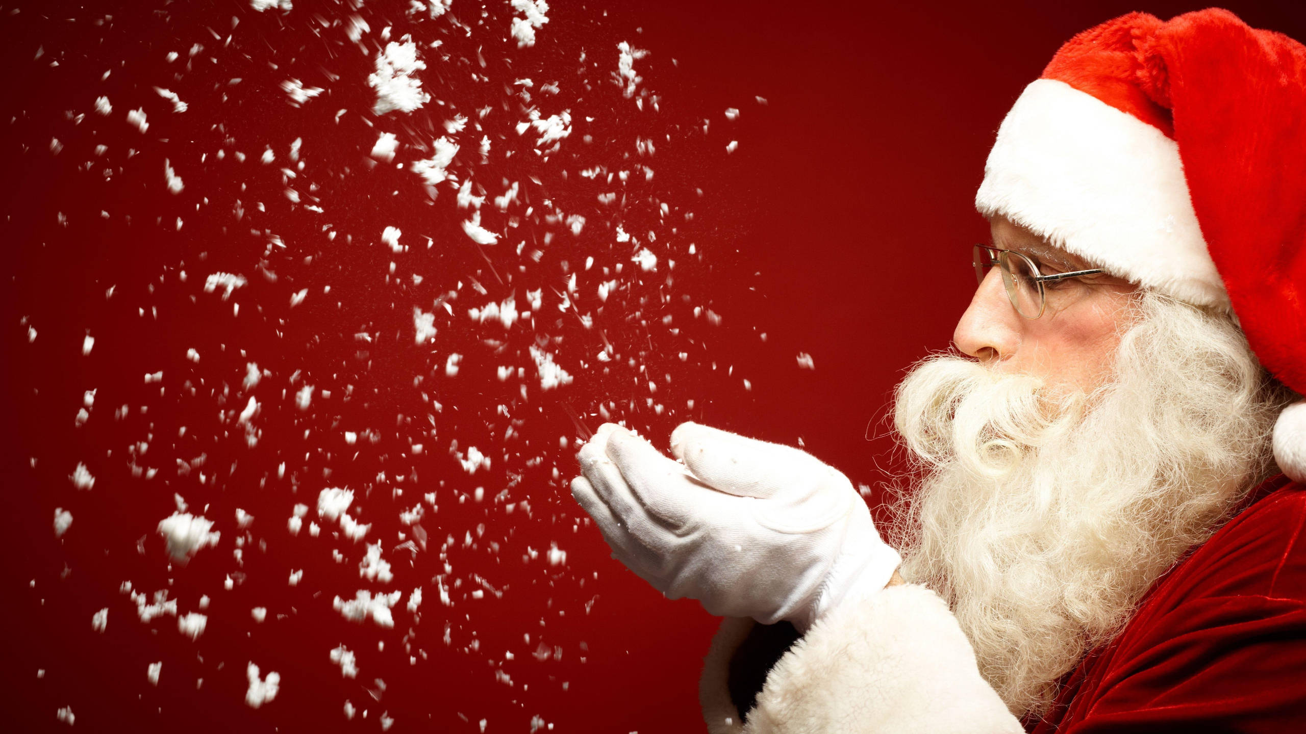 Santa Claus Blowing Snow 壁紙 — 聖誕老人從戴手套的手吹出雪花，襯托鮮豔的紅色背景。