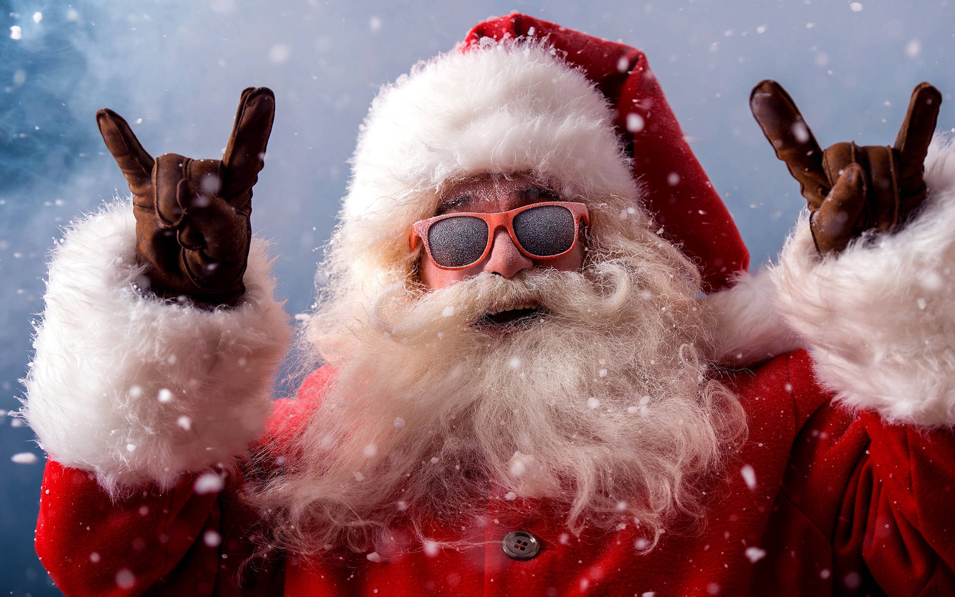 Santa Claus In Rock And Roll Gesture 壁紙 — 聖誕老人戴著紅色太陽眼鏡和毛邊外套，在藍天下飄雪中做出搖滾手勢。