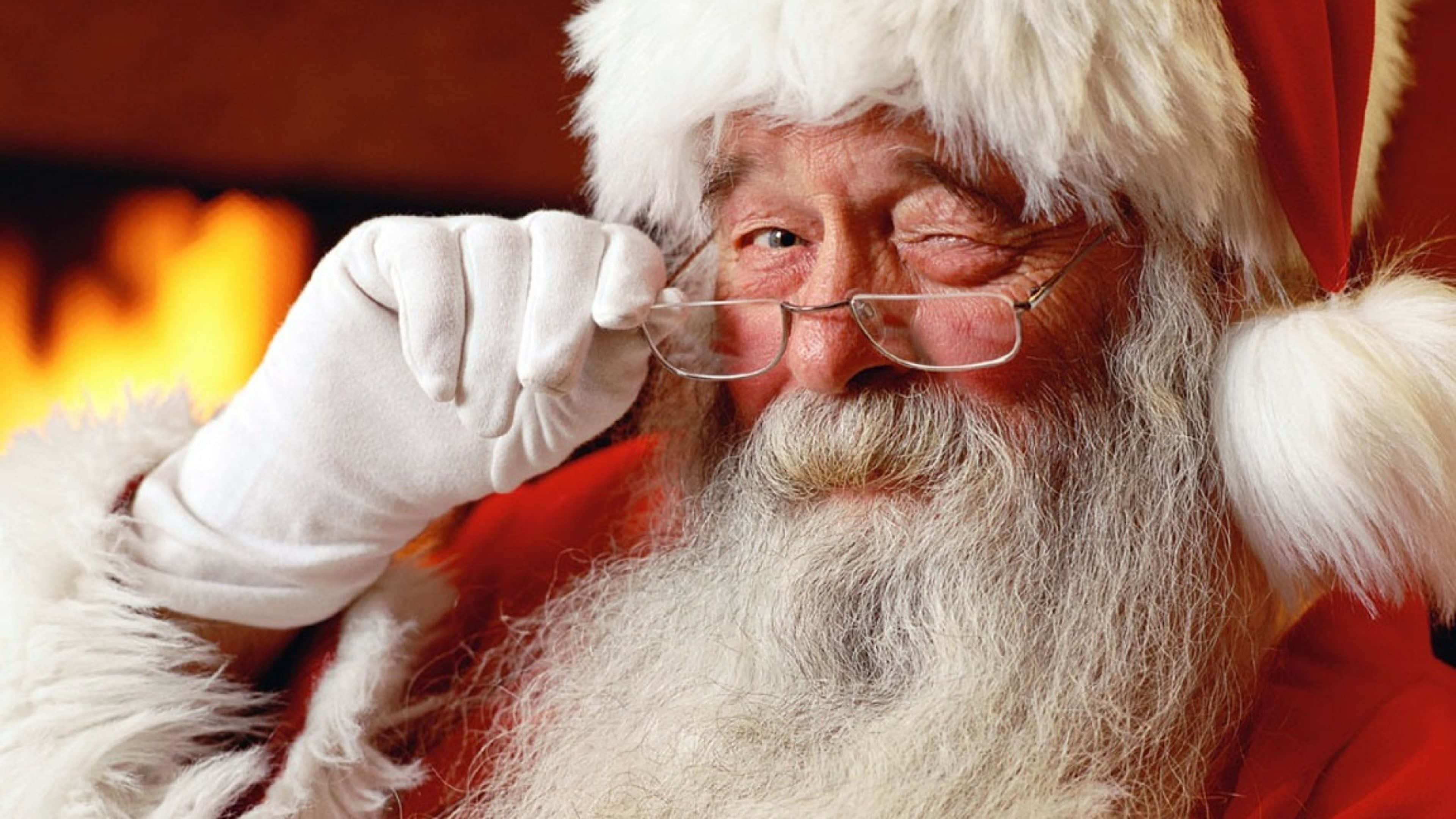 Santa Claus 桌布 壁紙 — 聖誕老人身穿紅色西裝和白色毛邊，在閃爍的壁爐旁微笑著調整眼鏡。