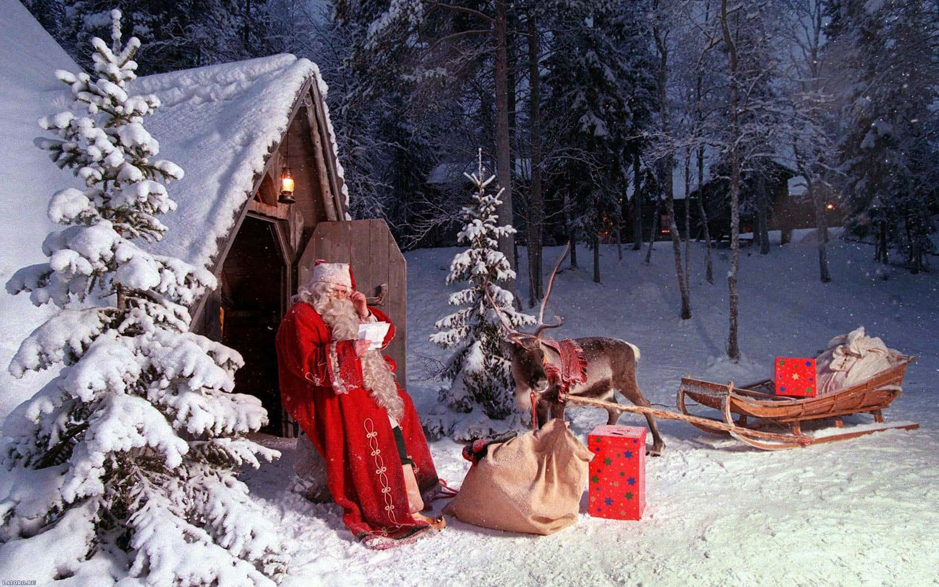 Santa Claus 桌布 壁紙 — 聖誕老人身穿紅色長袍，站在他的馴鹿和雪橇旁邊，在冬季森林中一間被雪覆蓋的小屋外。